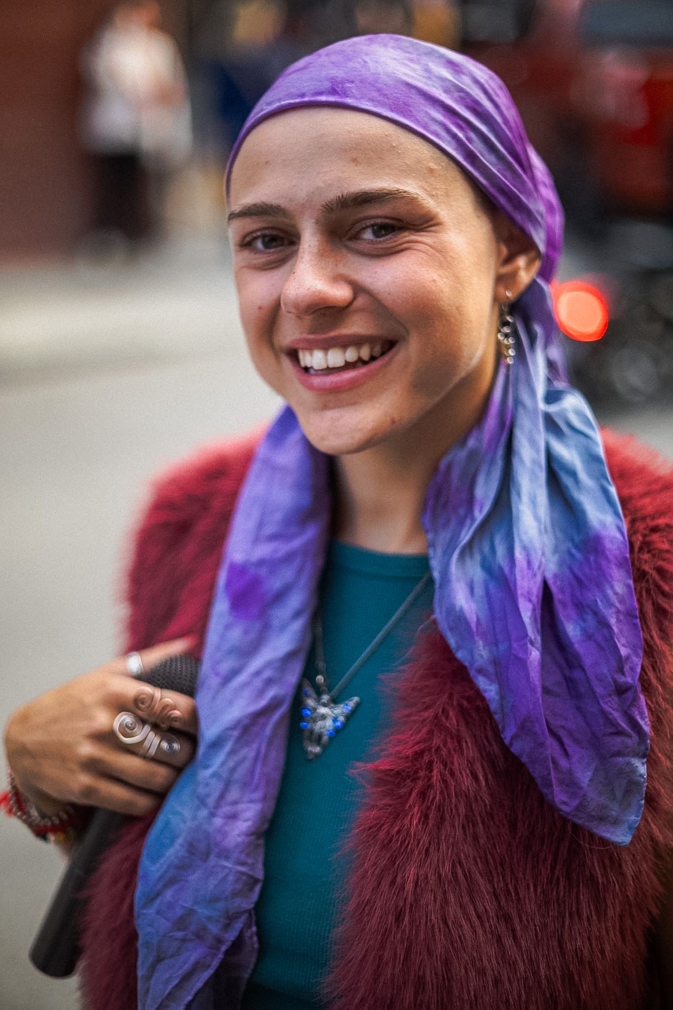 Smiling young woman wearing a purple head scarf, teal top, red fur vest, and butterfly necklace outdoors.
