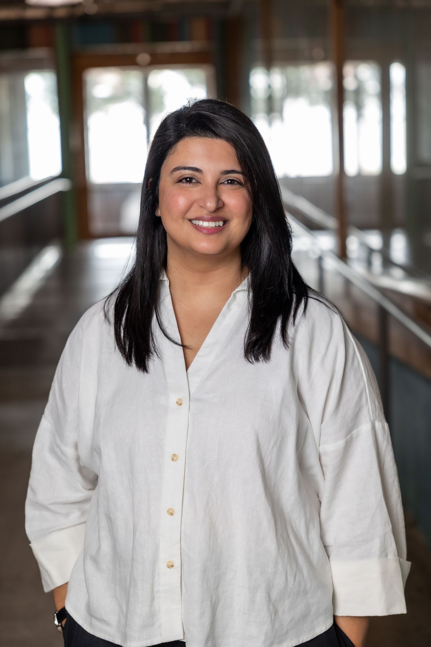 Woman with black hair in a white linen button-up shirt smiling in a modern indoor hallway.