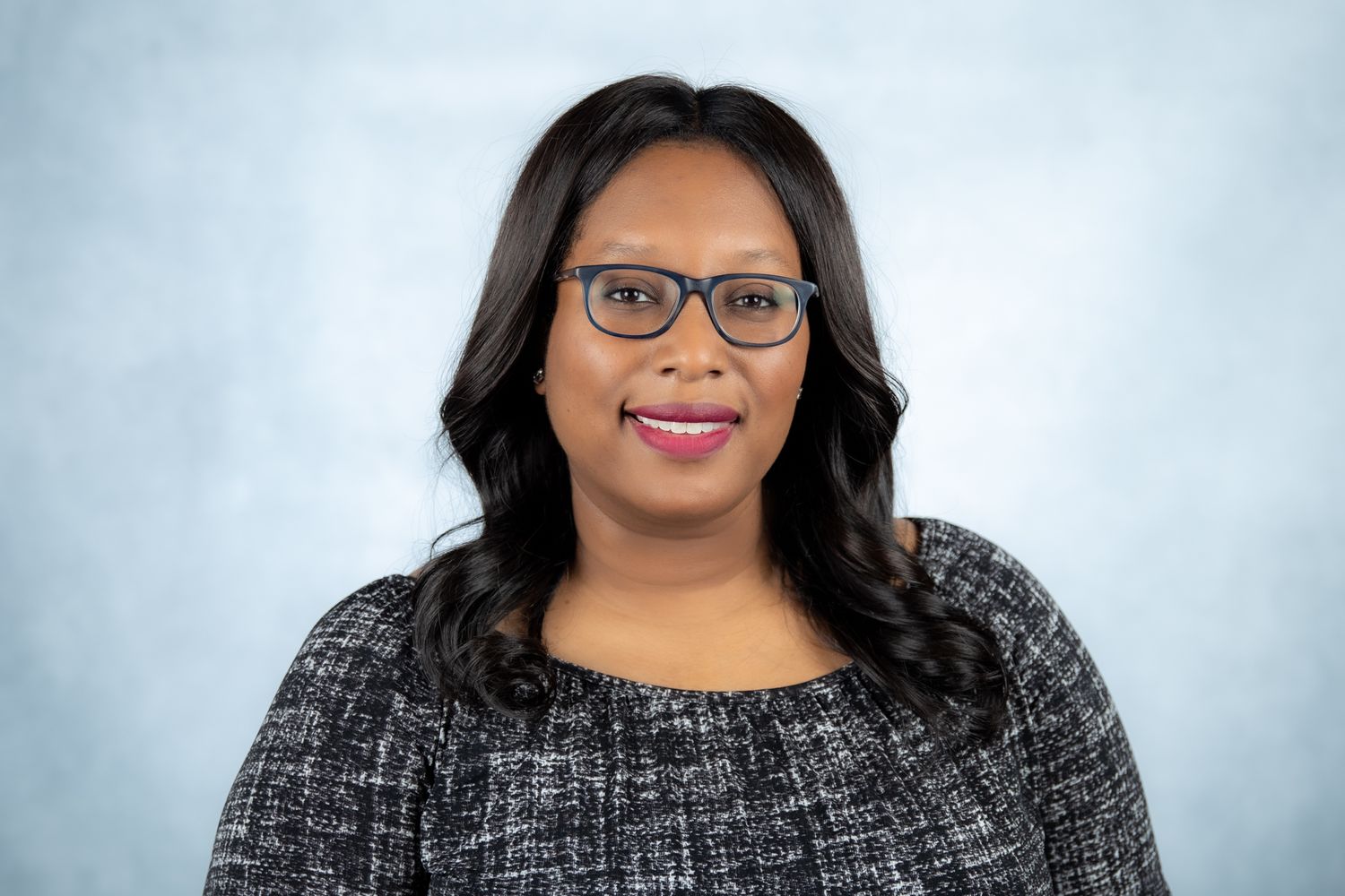Smiling woman with dark wavy hair wearing glasses and gray tweed top on blue background.