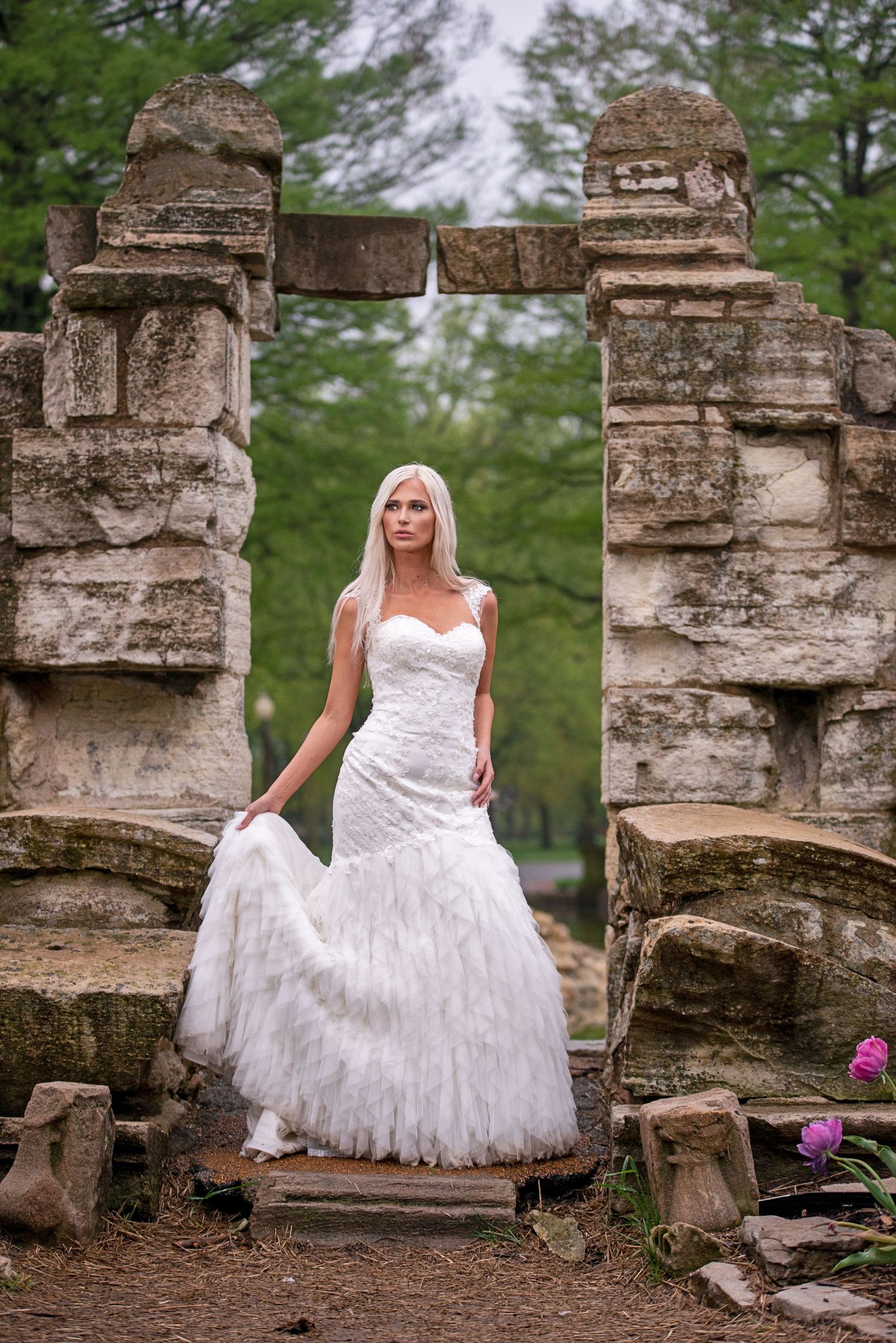 Blonde bride in white mermaid gown posing in ancient stone ruins surrounded by lush green trees.