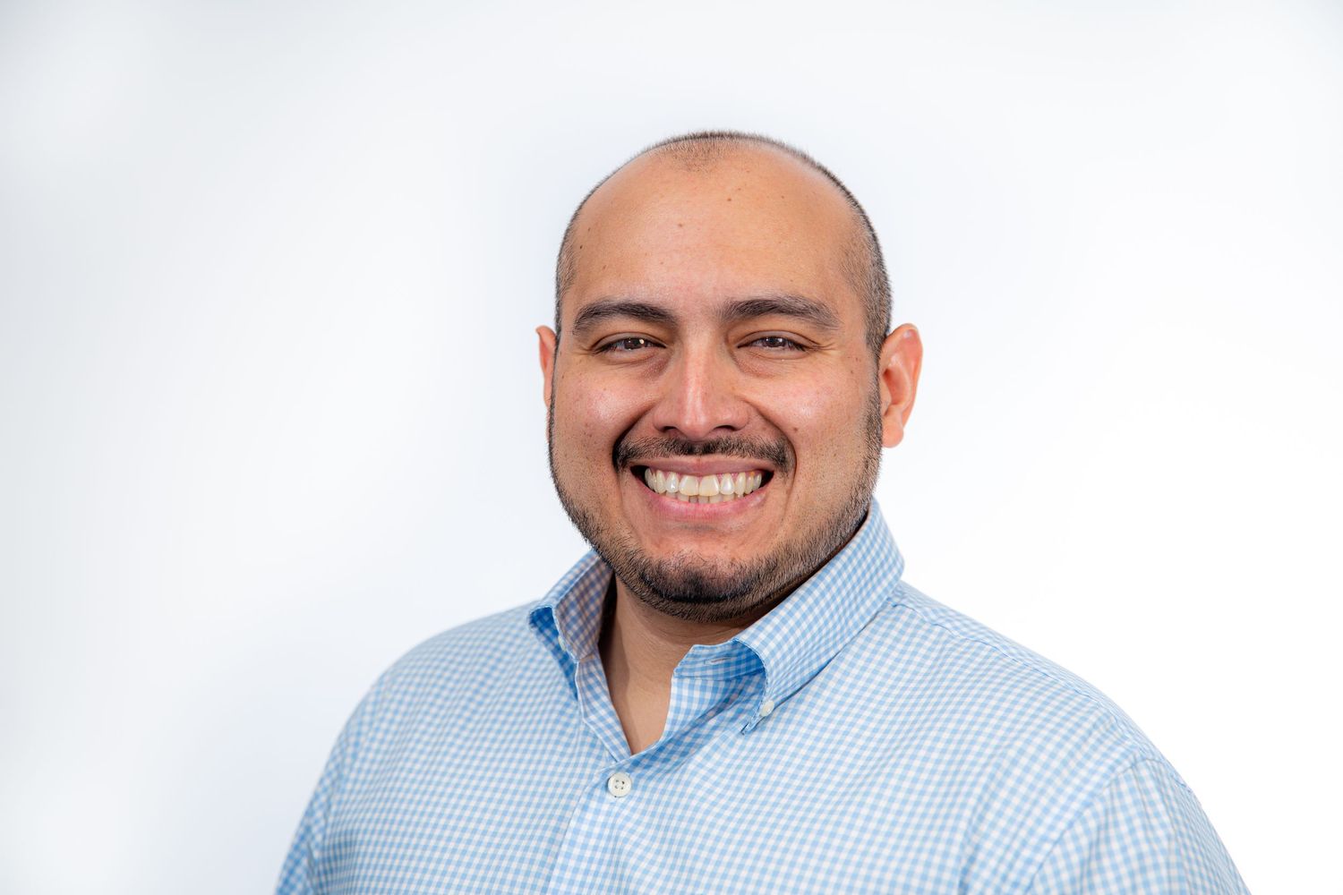 Smiling bald man with a beard wearing a light blue checkered shirt against a white background.