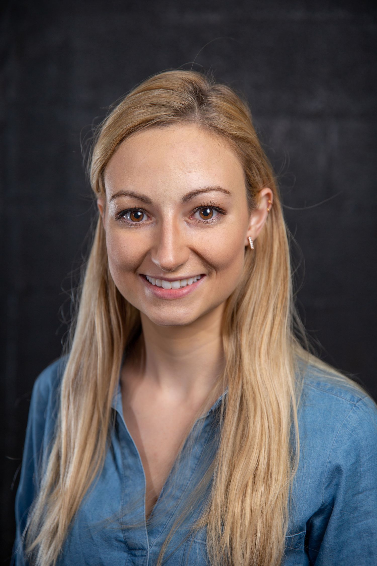 Smiling blonde woman with long hair wearing a blue denim shirt against a dark background.