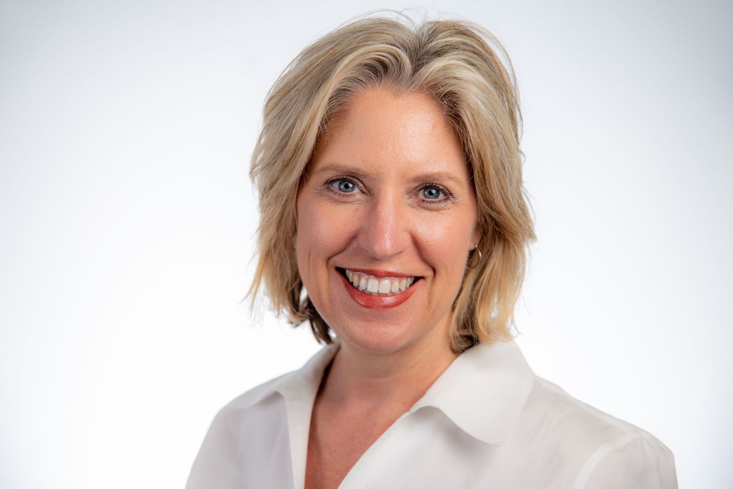 Smiling blonde woman with short wavy hair wearing a white blouse against a light background.