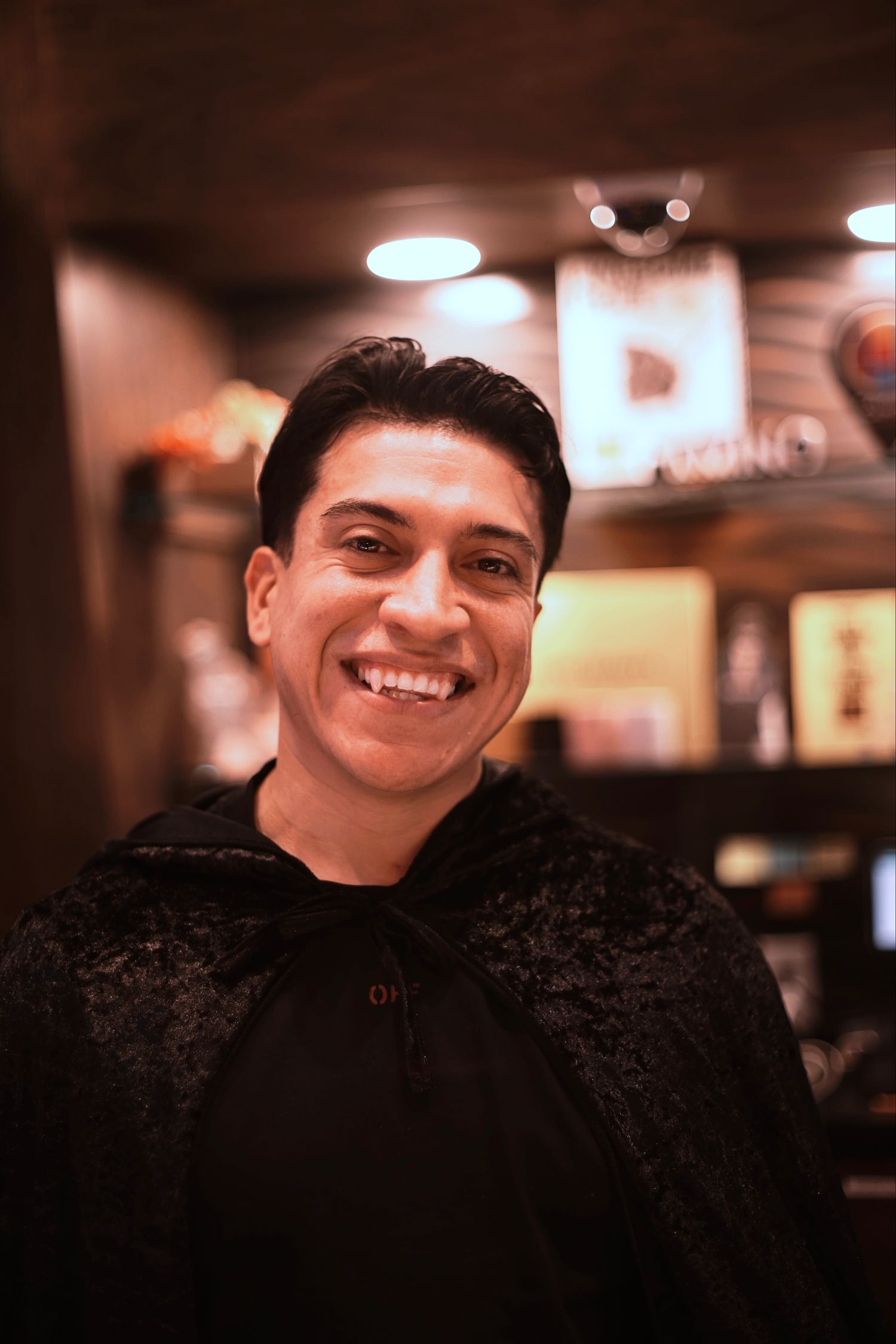 Smiling young man in black cape costume posing indoors at warmly lit venue.