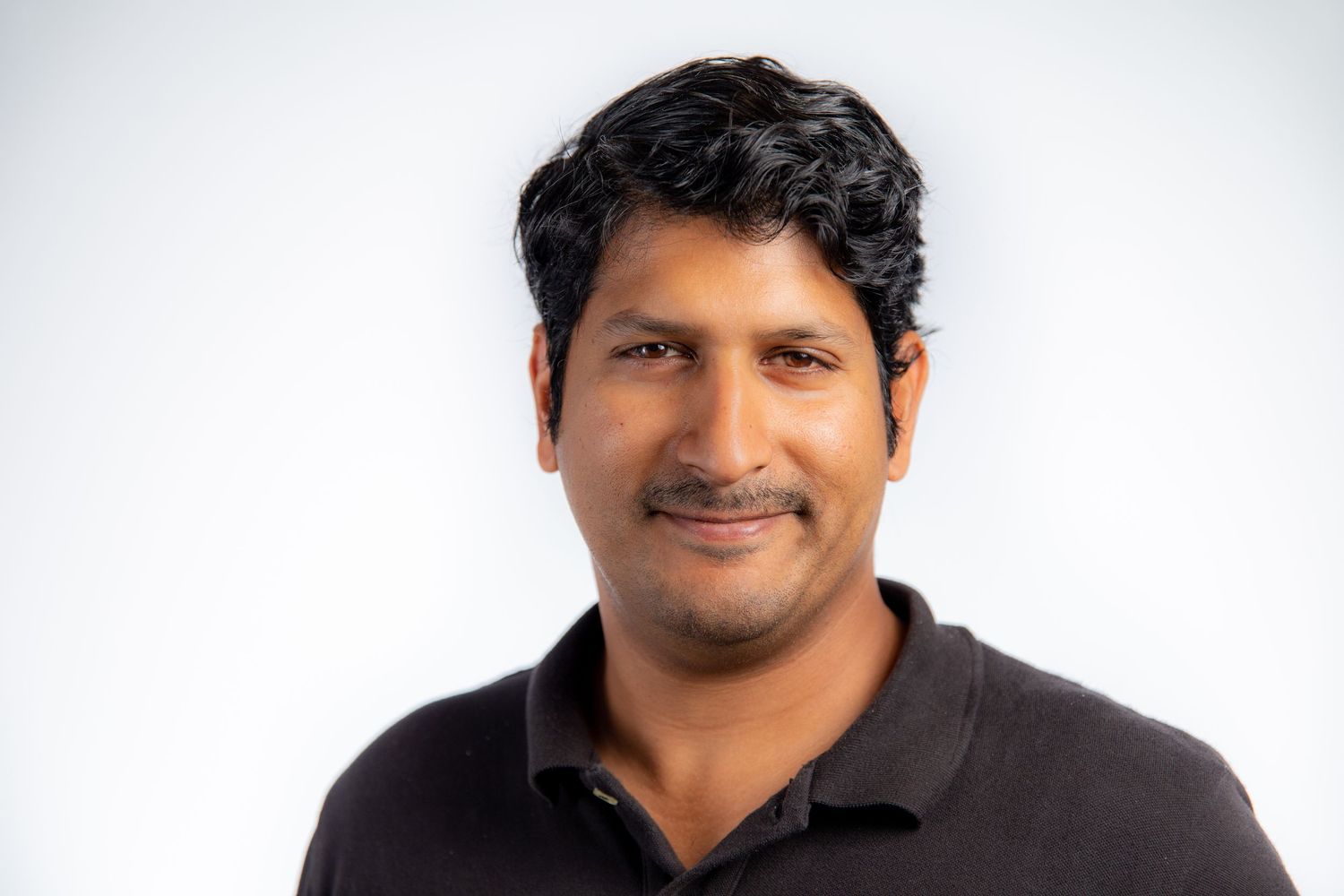 Professional headshot of a man in a dark polo shirt smiling against a light gray background.