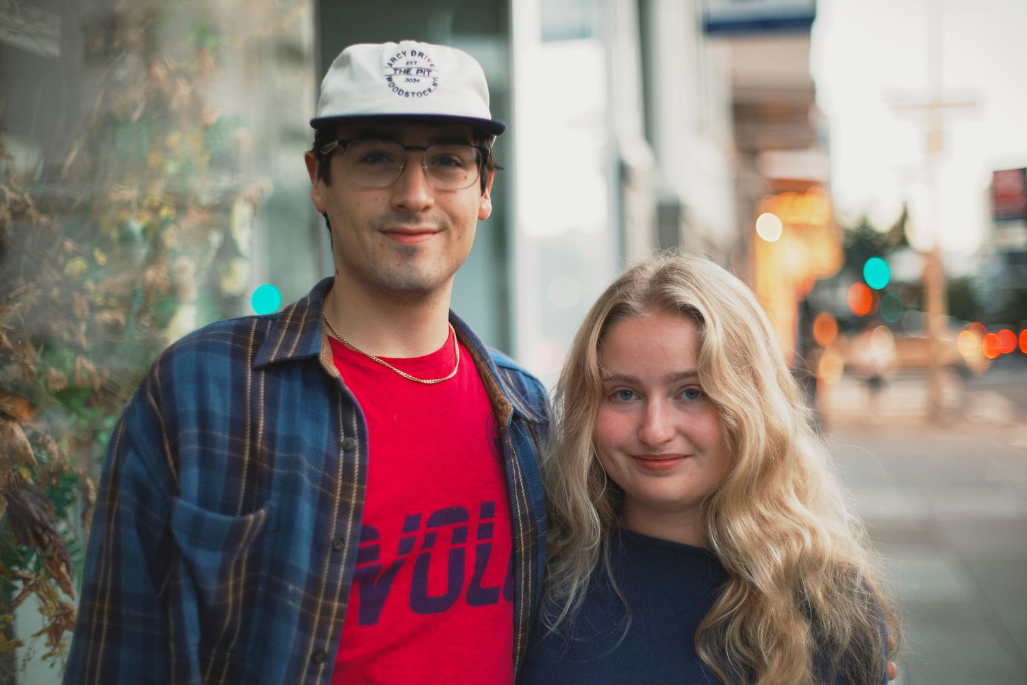 Young couple smiling outdoors, man in plaid shirt and cap, woman with long blonde hair on city street.