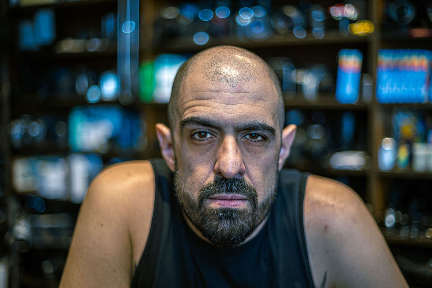 Bald man with beard and intense expression sits in a dimly lit room with shelves behind him.
