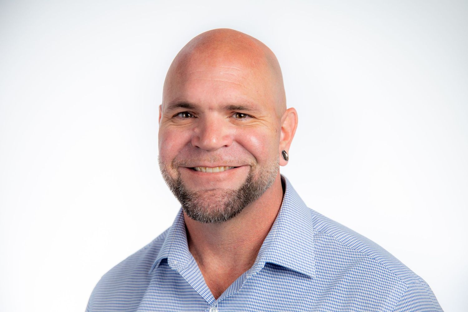 Smiling bald man with a goatee wearing a light blue striped shirt against a white background.