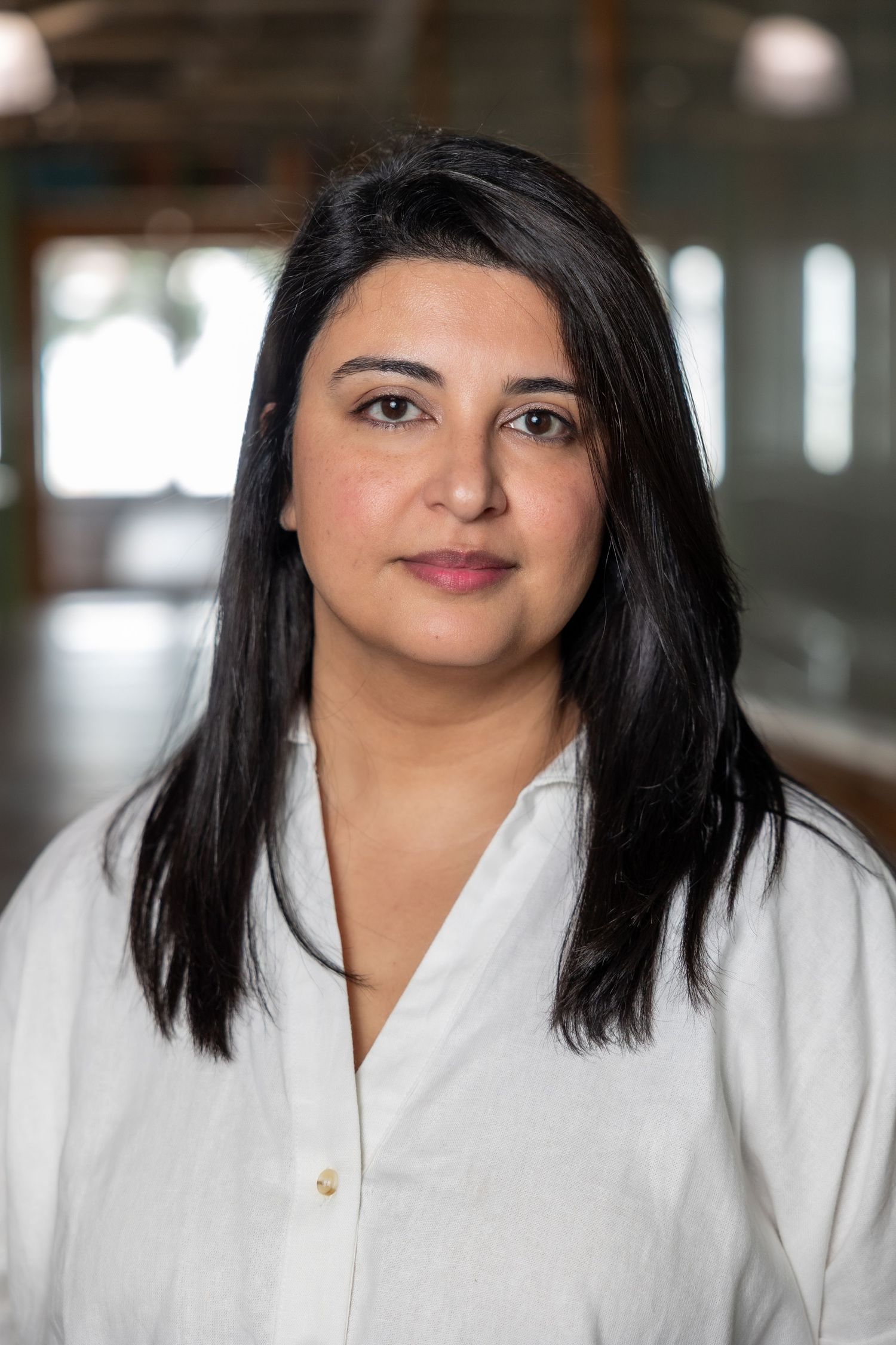 Professional woman with black hair in a white linen shirt posing in a modern office environment.