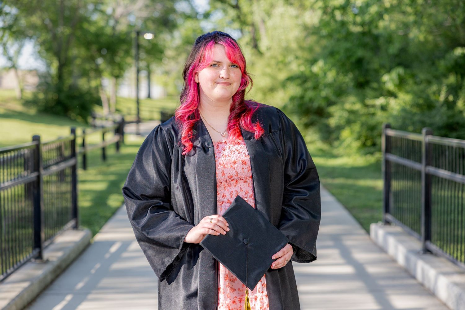 A smiling graduate with pink hair holds cap wearing black gown on a sunny outdoor walkway.