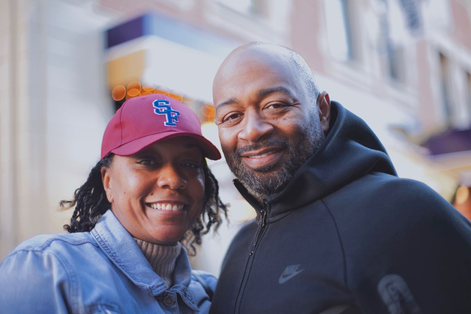 Smiling couple poses together outdoors, woman wearing red SF baseball cap and denim jacket.