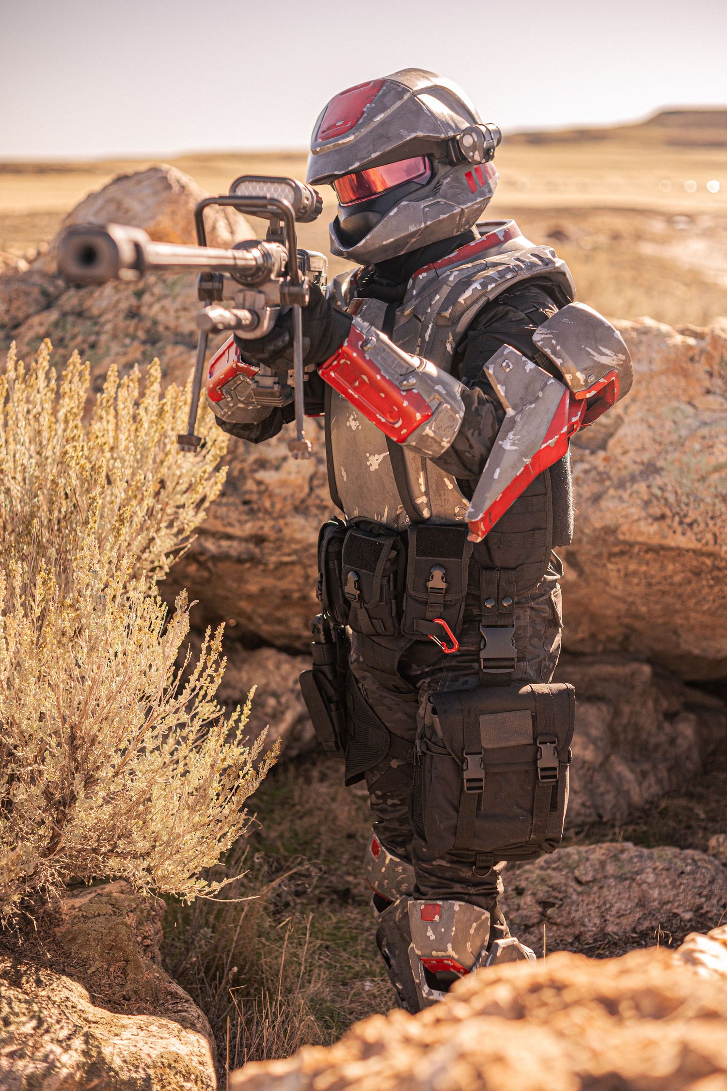 Cosplayer in red and grey sci-fi armor aiming futuristic rifle from behind desert rocks.