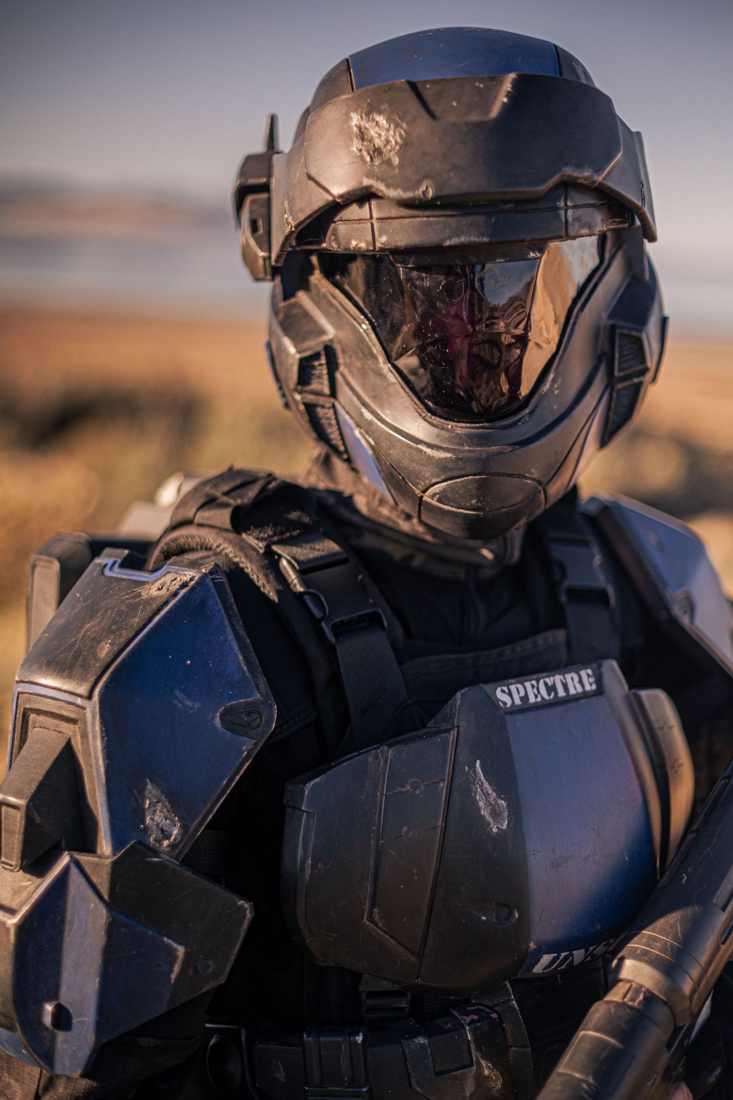 Close-up portrait of a person wearing weathered black futuristic armor with SPECTRE chest plate insignia.