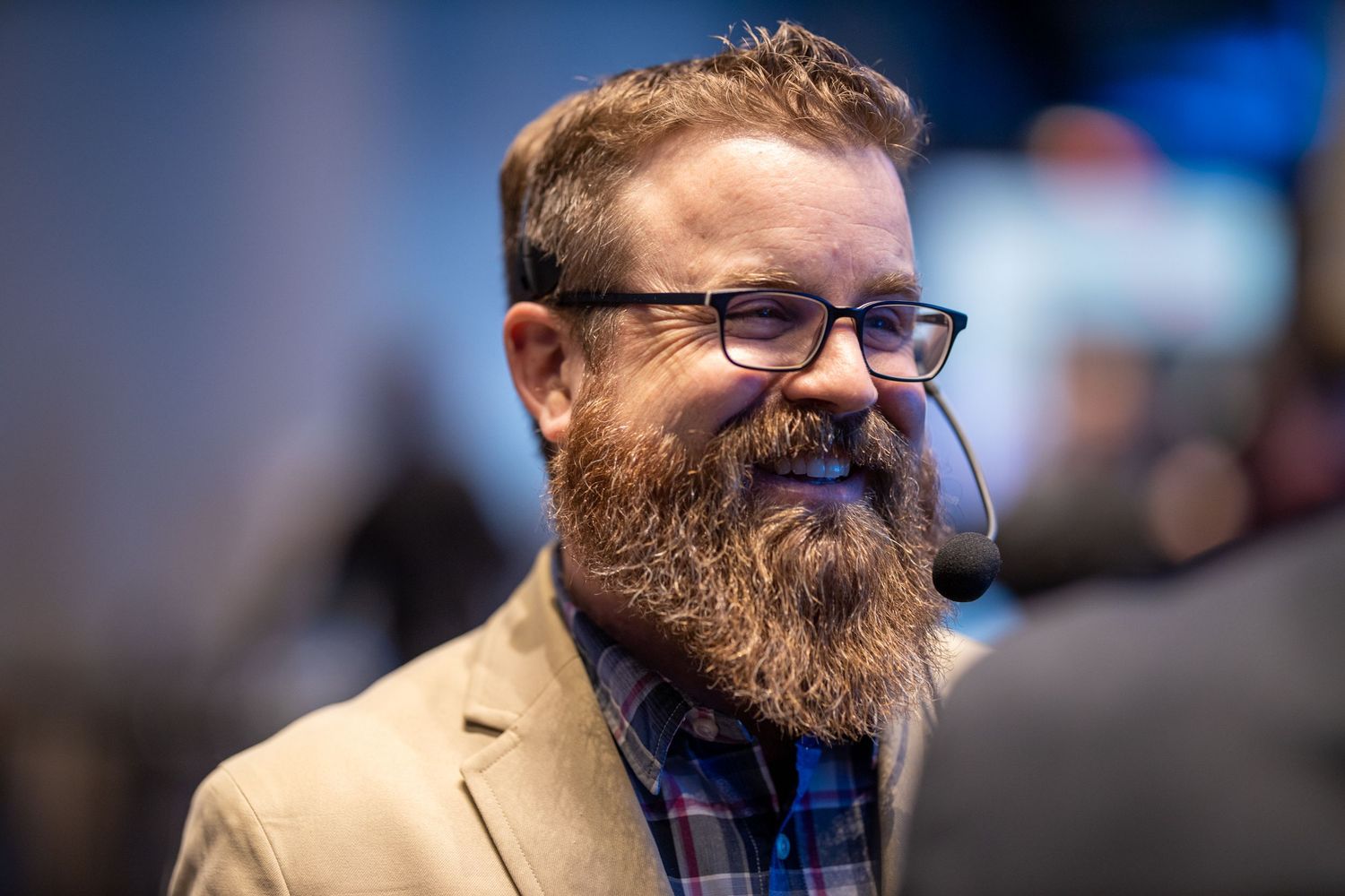 Smiling bearded man with glasses and headset mic wearing a plaid shirt and tan blazer at an event.