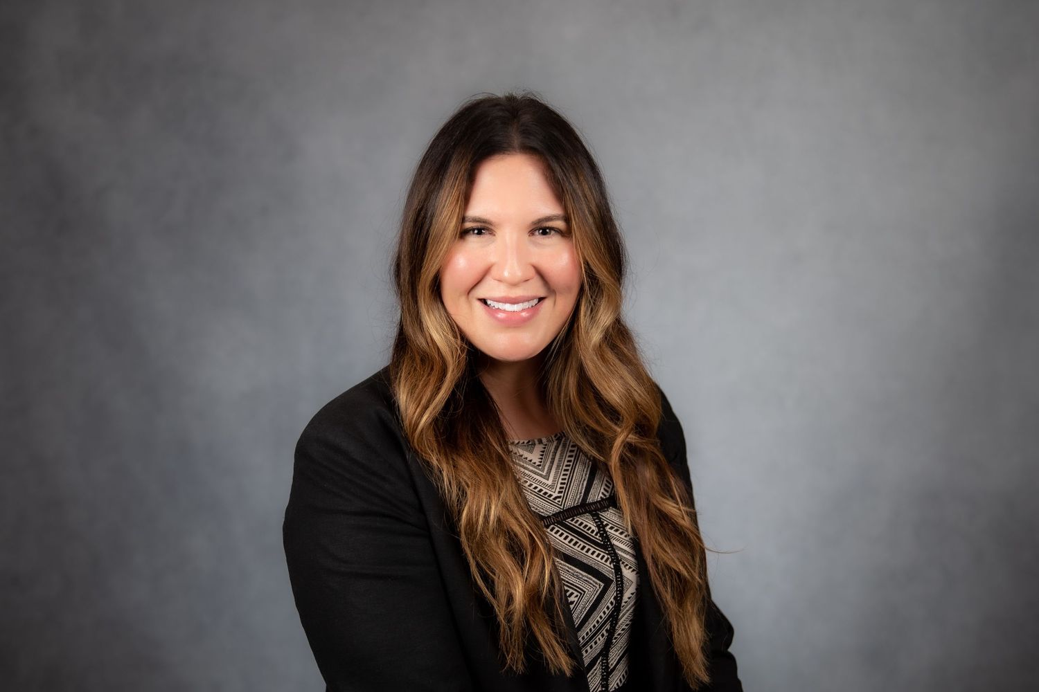 Professional woman with long brown hair wearing a black blazer on gray background.