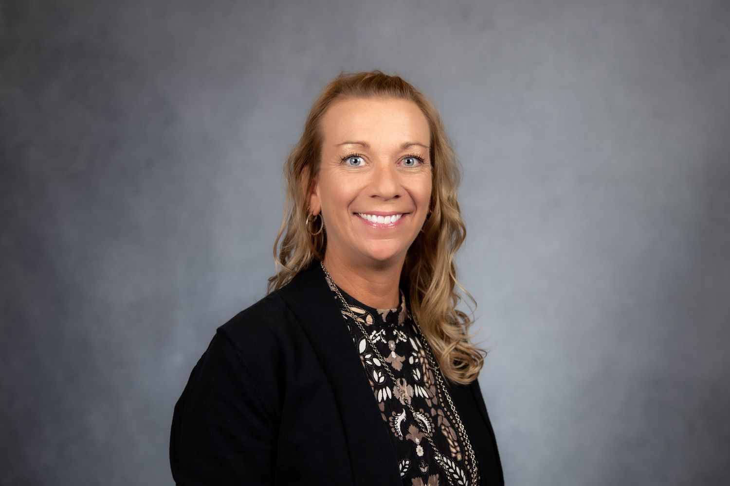 Smiling blonde woman in black blazer and patterned top posing on gray background.