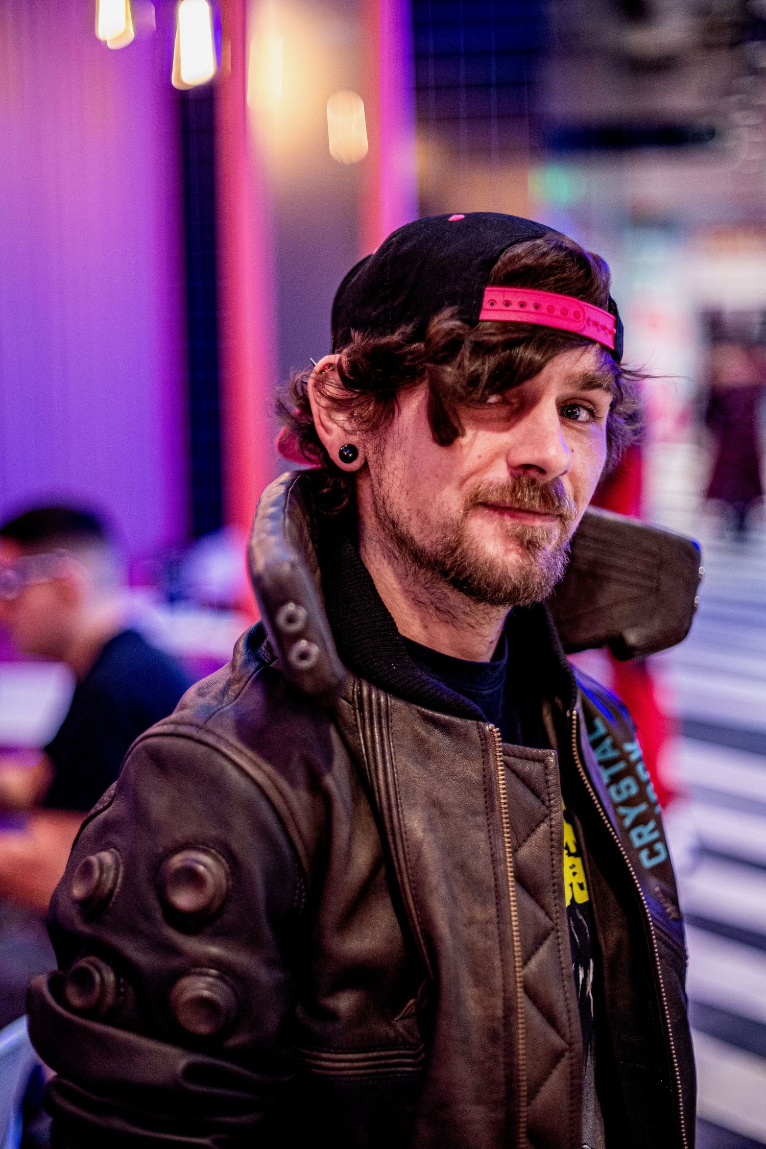 Man in black cap and leather jacket with cyberpunk accessories poses under purple neon lights.