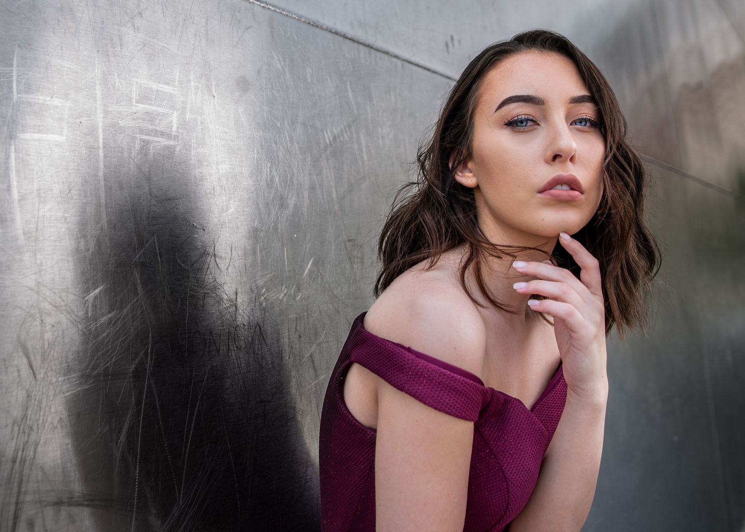 Young brunette woman in burgundy off-shoulder dress posing against a metallic silver wall.