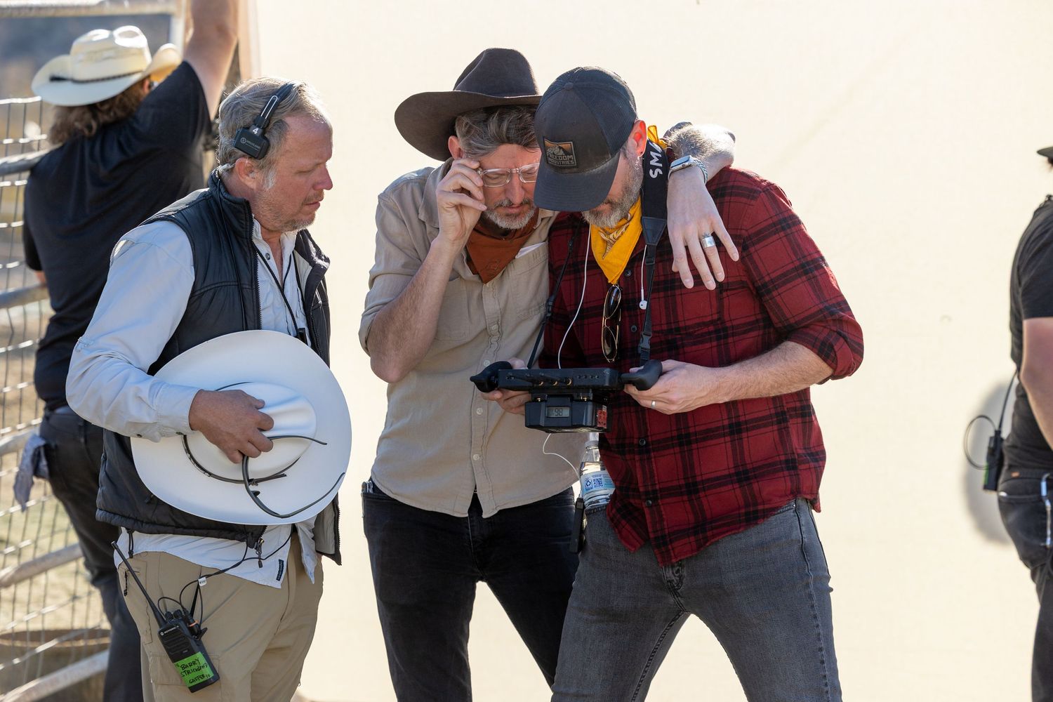 Three men on a film set review footage on a camera monitor outdoors in bright sunlight.