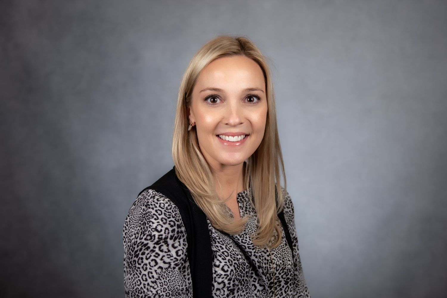 Blonde woman smiling in a leopard print blouse on a gray background.