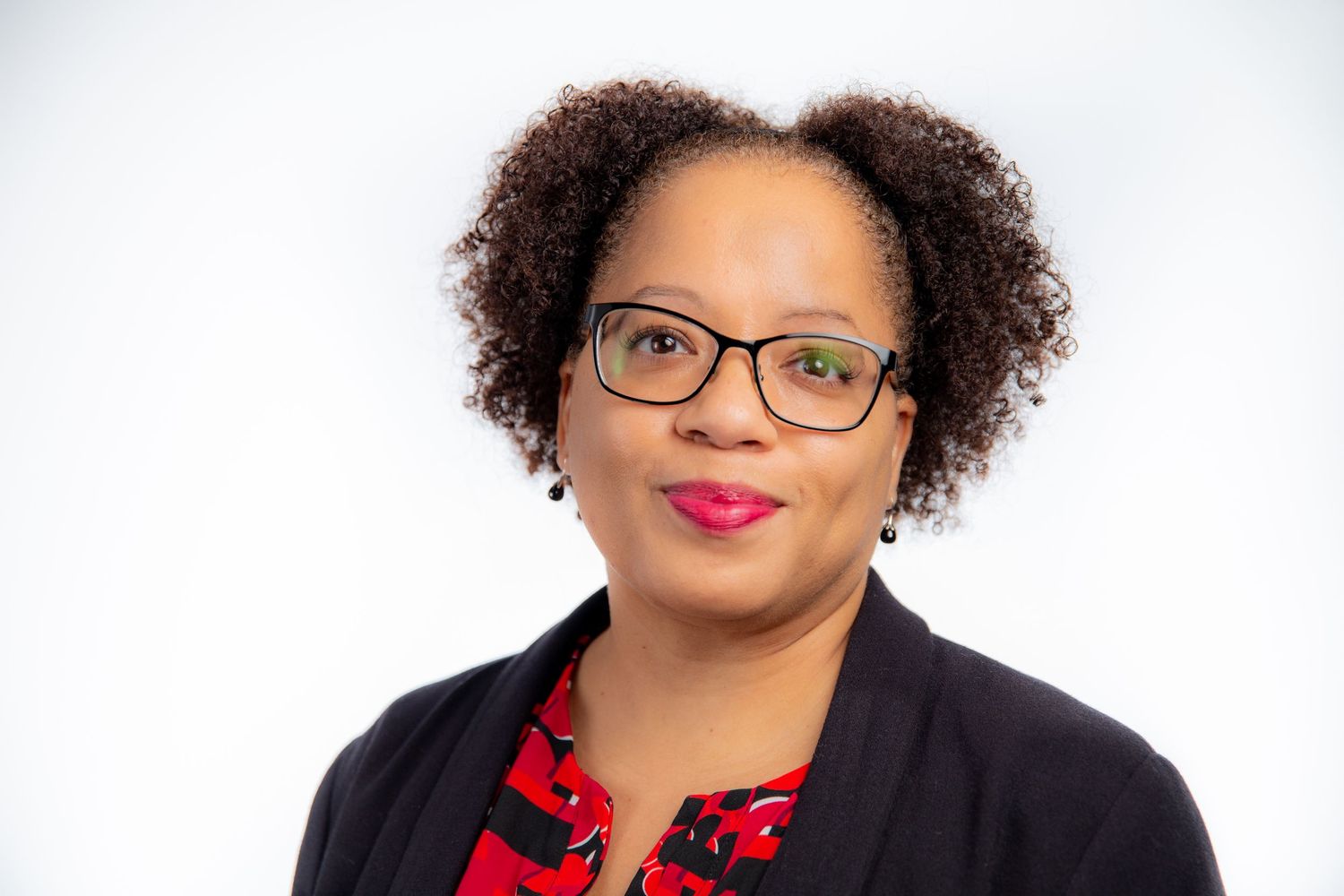 Professional woman with curly hair wearing glasses and a red floral top with a black blazer.