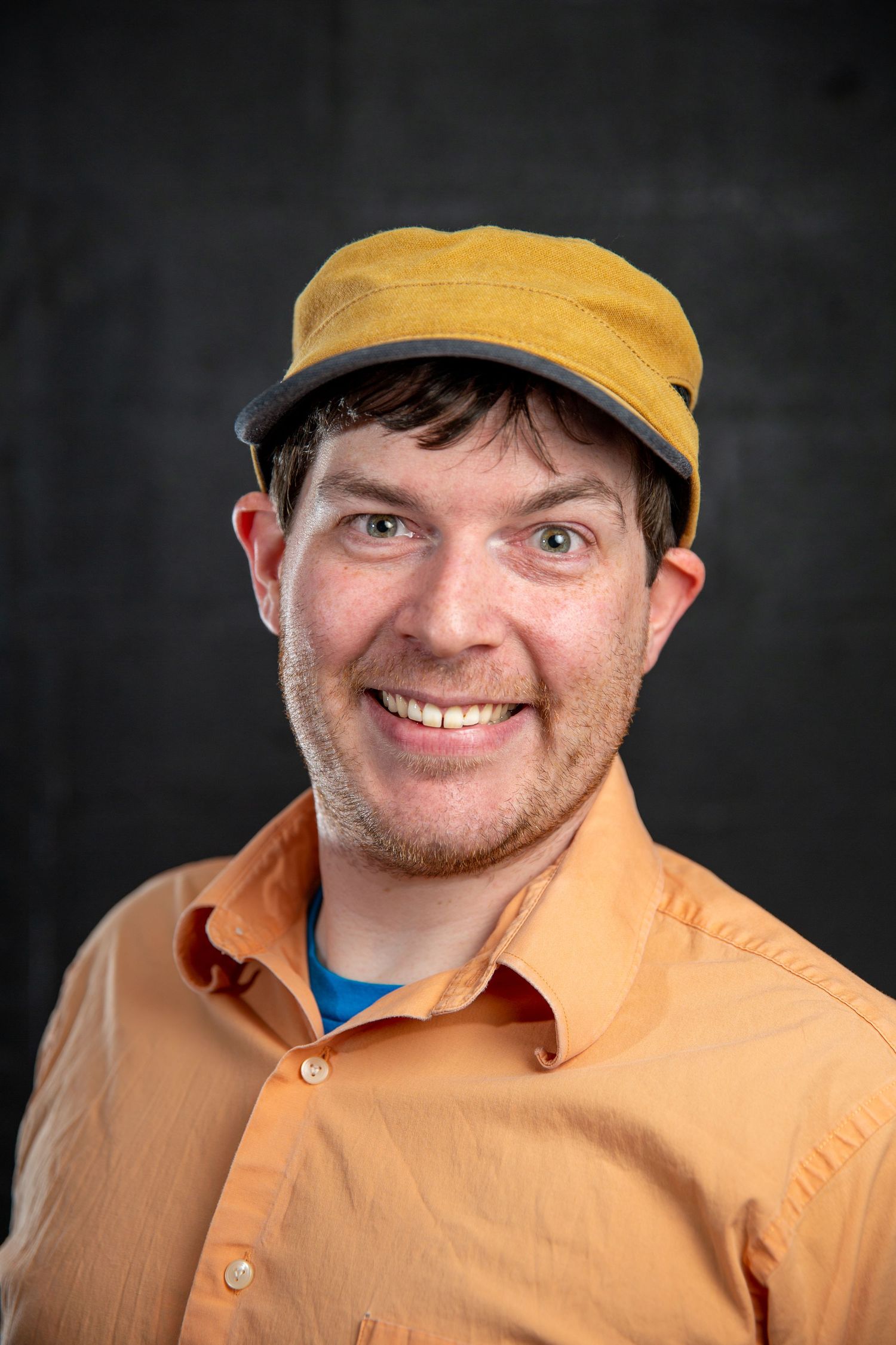 Smiling man wearing an orange button-up shirt and yellow flat cap poses for a professional headshot.