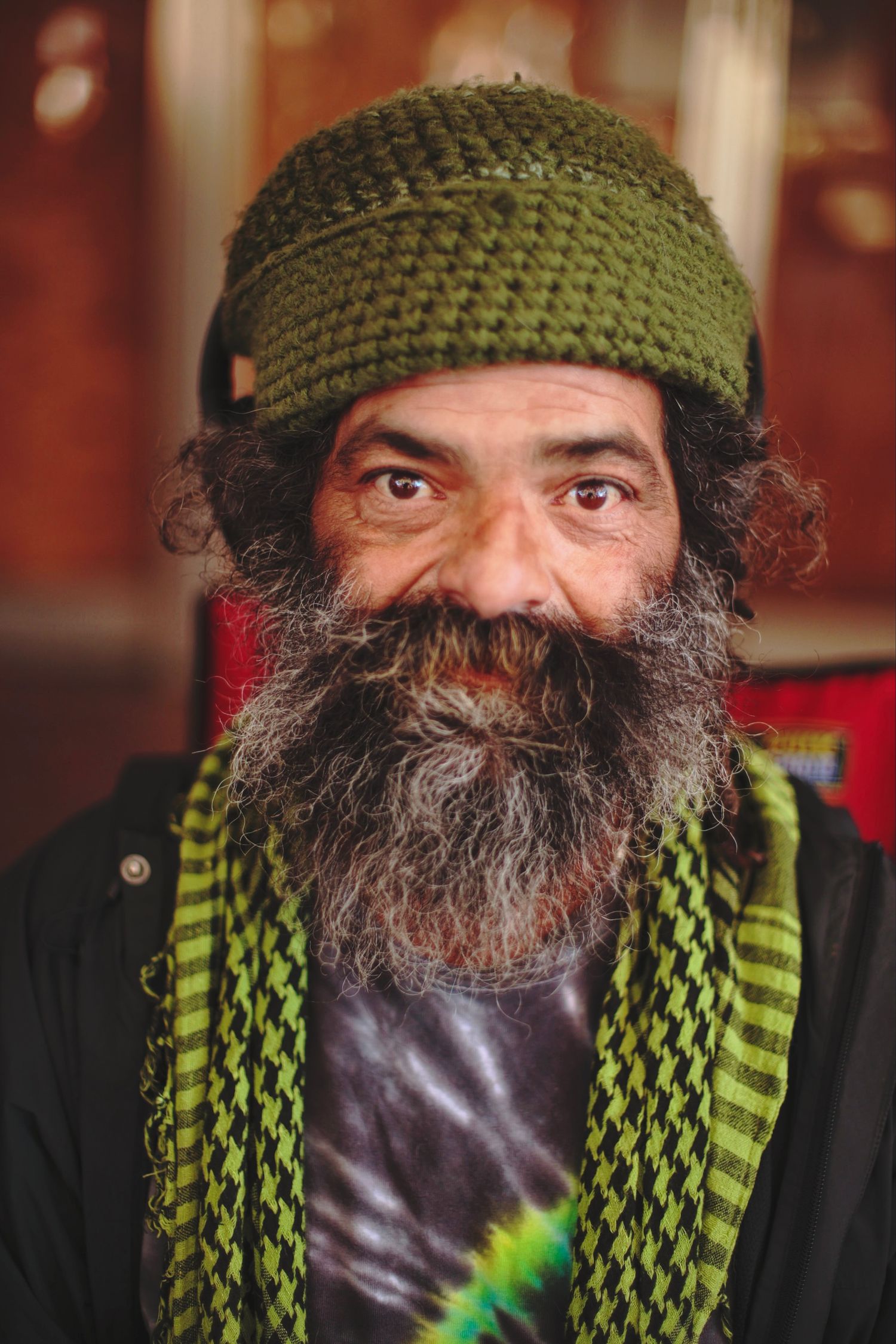Older man with a long gray beard wearing a green knit hat and striped scarf smiling at camera.