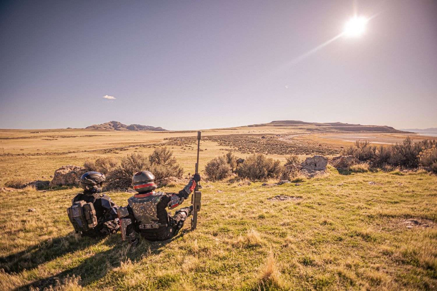 Two armored soldiers lying in green grassland aiming rifles toward distant rocky hills.