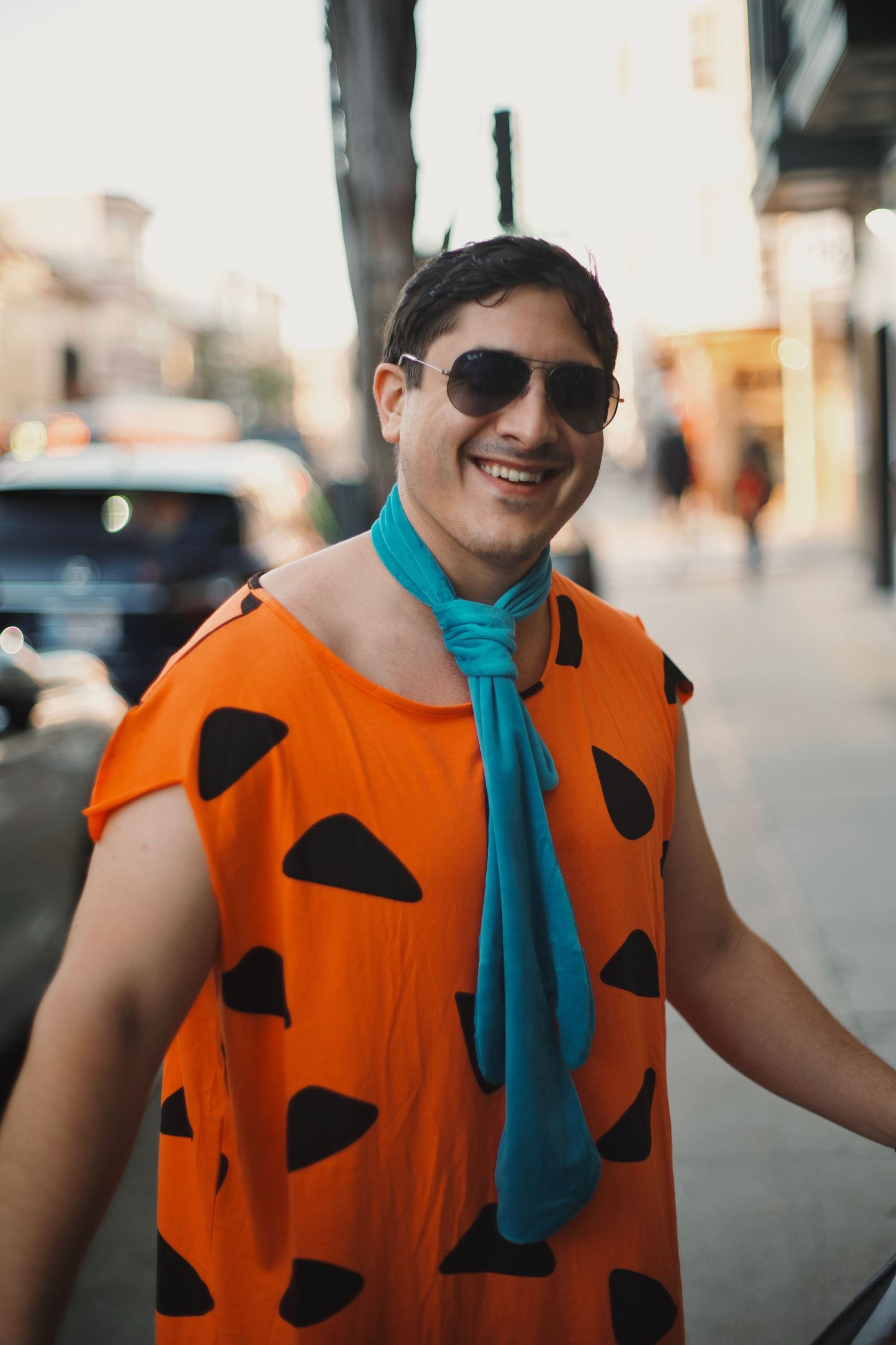 Smiling man in Fred Flintstone Halloween costume with sunglasses on a city street.