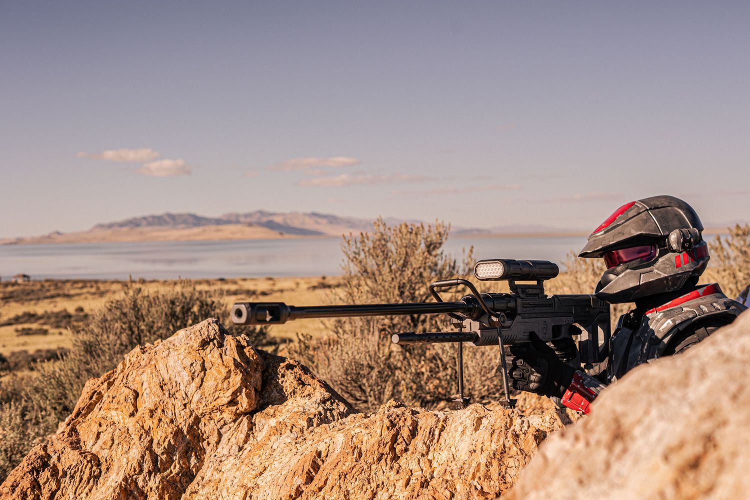 A cosplayer in a red helmet aims a sniper rifle from rocky terrain with a scenic lake and mountains in the distance.