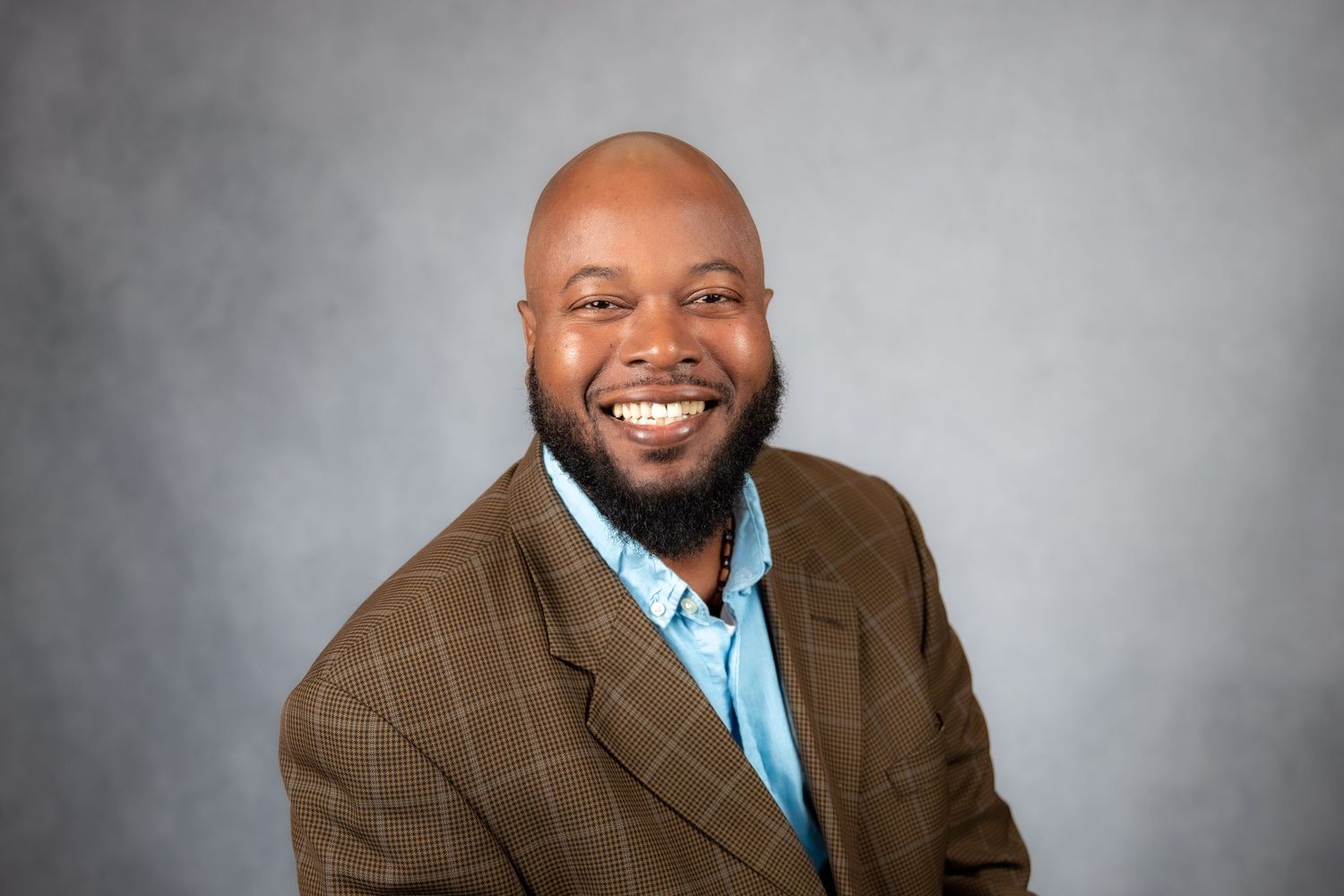 Smiling bald Black man with beard wearing brown plaid blazer and blue shirt against gray background.