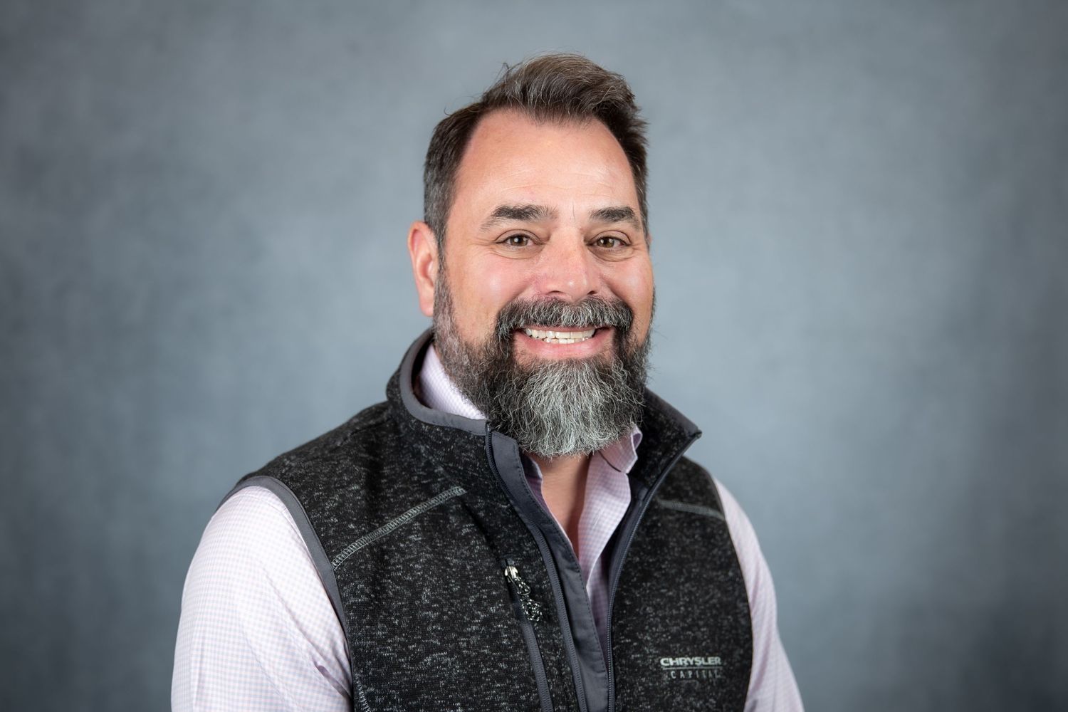 Bearded man smiling in a gray vest and white shirt on gray background.