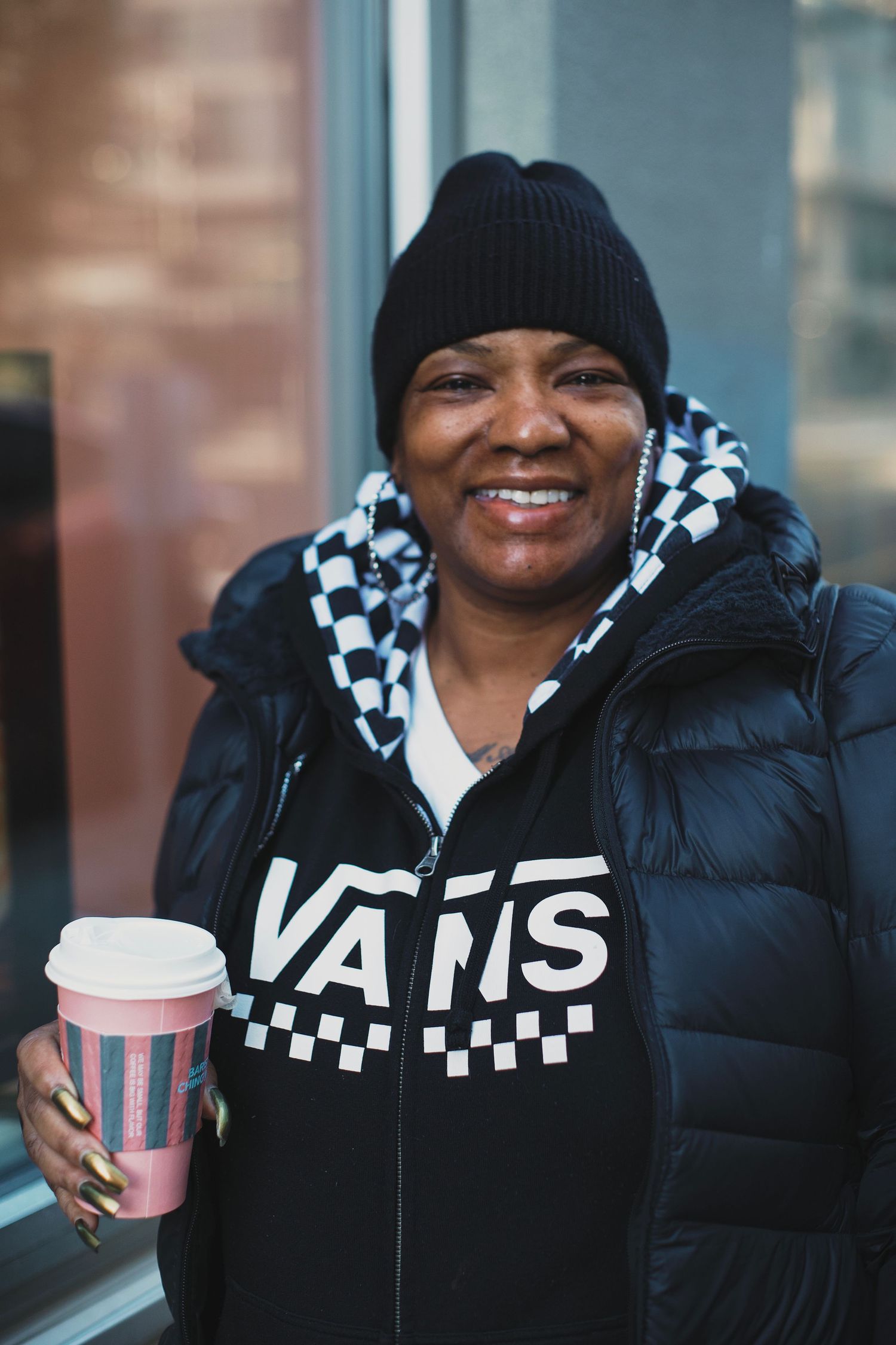 Smiling woman wearing a Vans hoodie and black beanie holding a pink coffee cup outdoors.