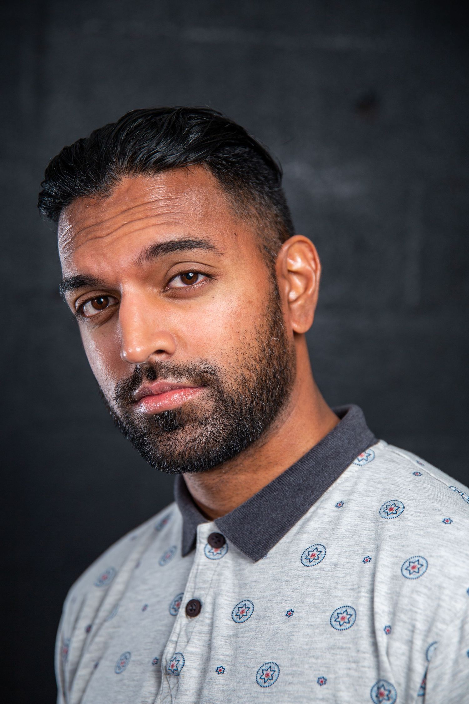 Close-up portrait of bearded man in gray patterned polo shirt against dark brick wall background.