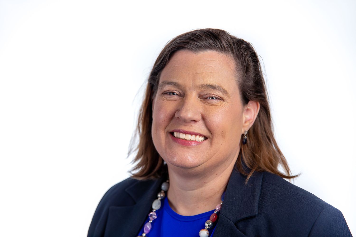 Professional headshot of a smiling woman with auburn hair wearing a blue top and navy blazer on white background.