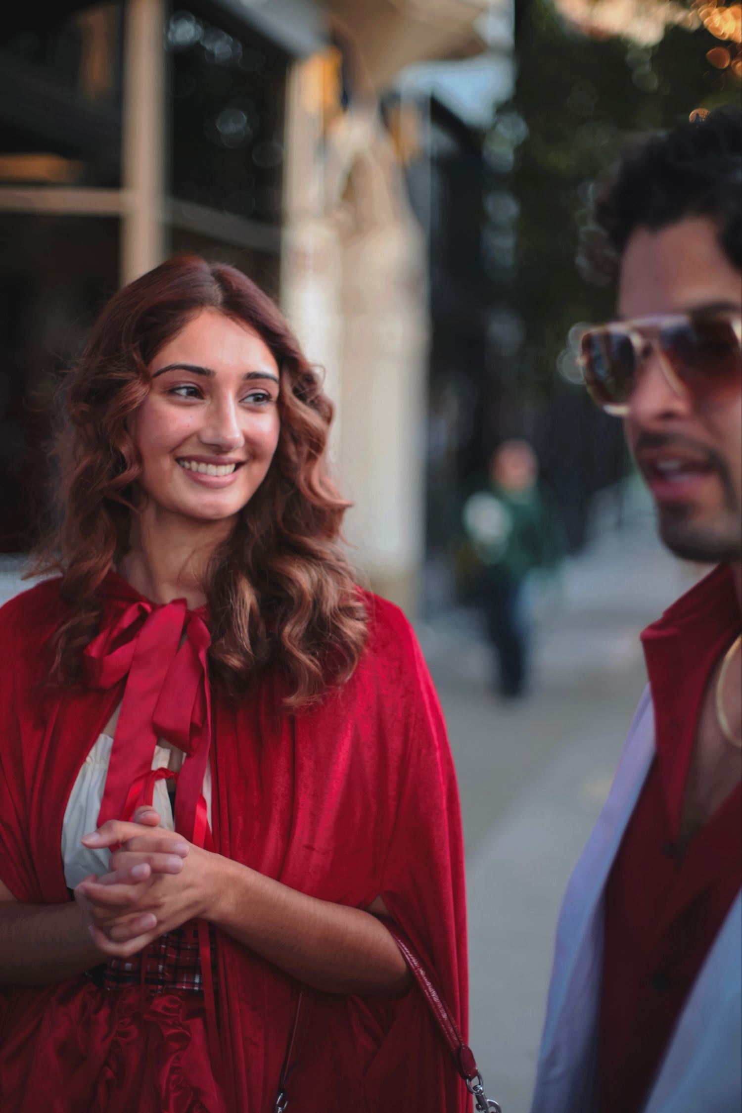 Smiling woman in red cape holding a glass on a city street at dusk.