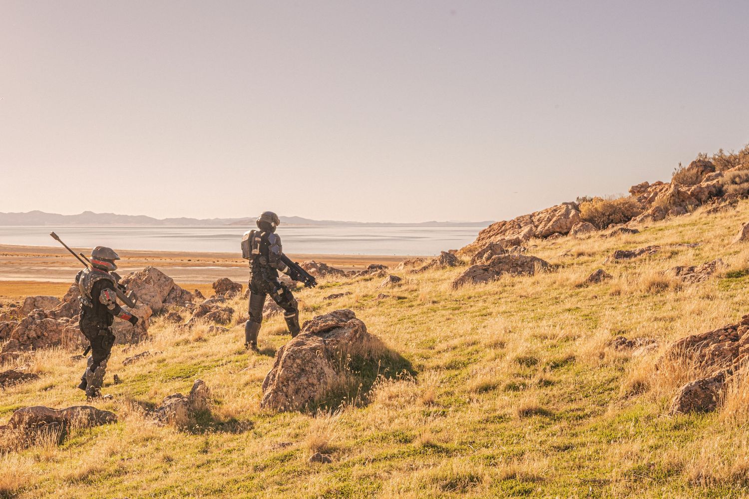 Two armed soldiers in black tactical gear patrolling a rocky grassy hillside with ocean views.