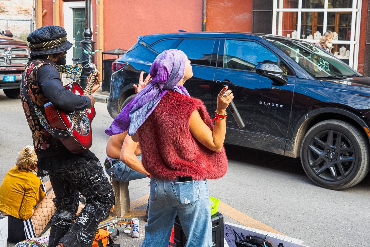 Two women photograph a blue SUV on a city street, one wearing a purple headscarf and red vest.