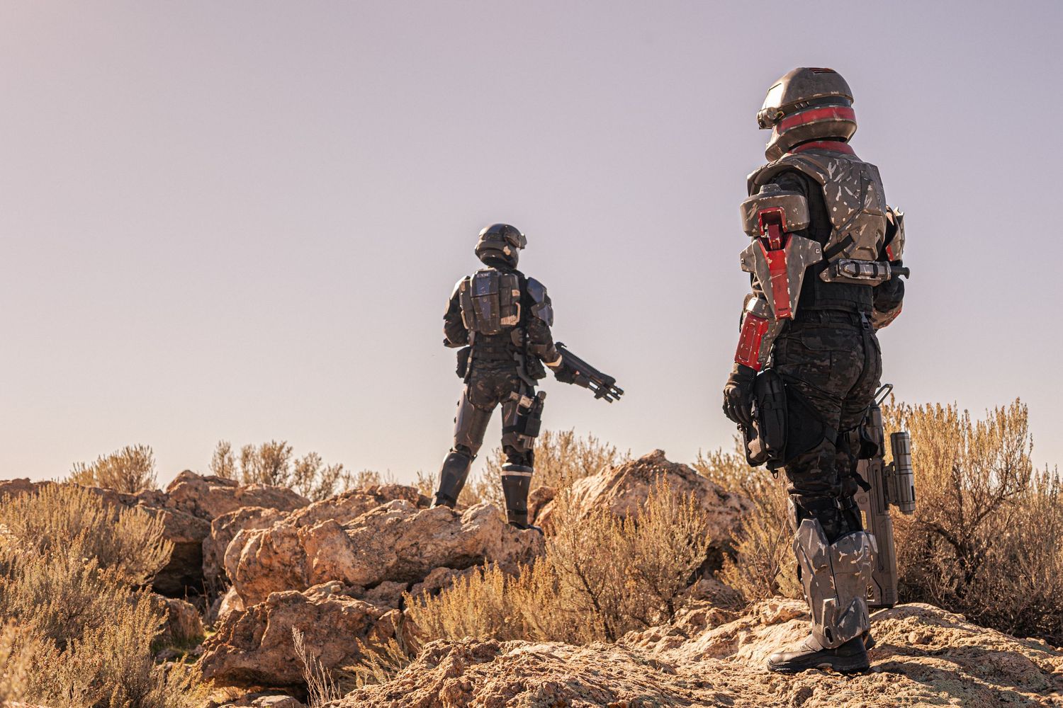 Two people in military sci-fi cosplay armor stand on rocky desert terrain under a clear sky.