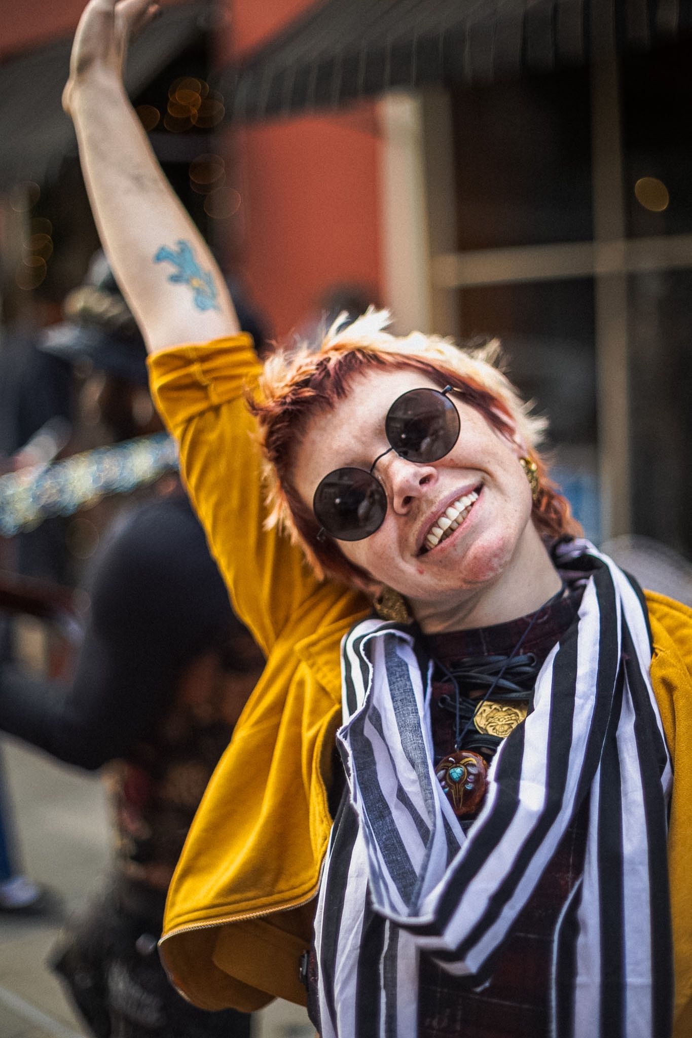 Happy woman in round sunglasses, yellow jacket, and striped scarf raises her arm showing a blue tattoo.