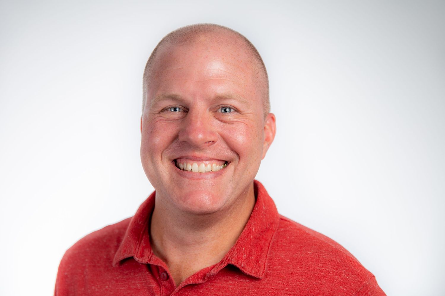 Smiling bald man wearing a red polo shirt poses for a professional headshot against a light gray background.