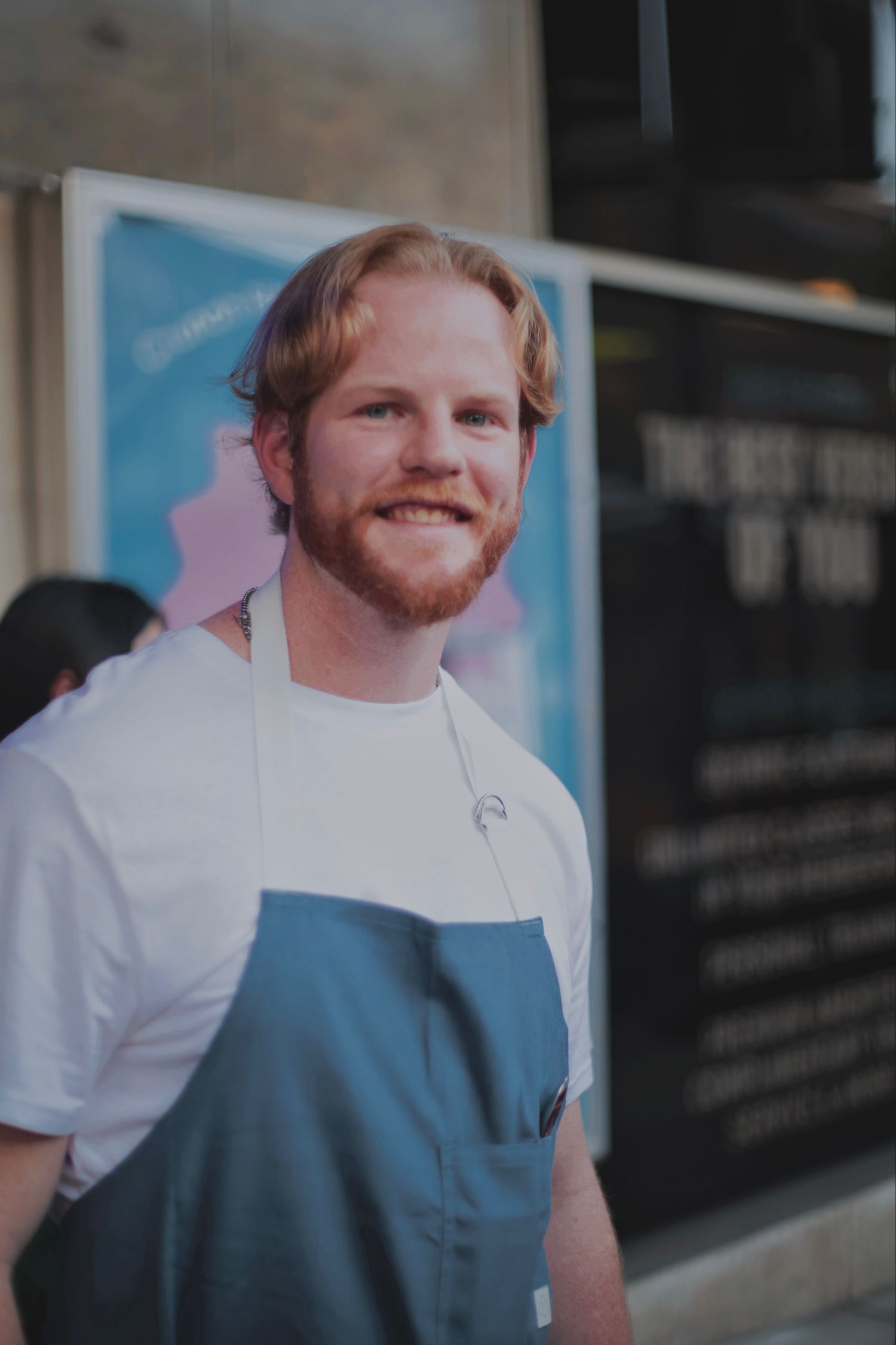 Cheerful redhead man with beard wearing a blue apron standing outdoors at an event.