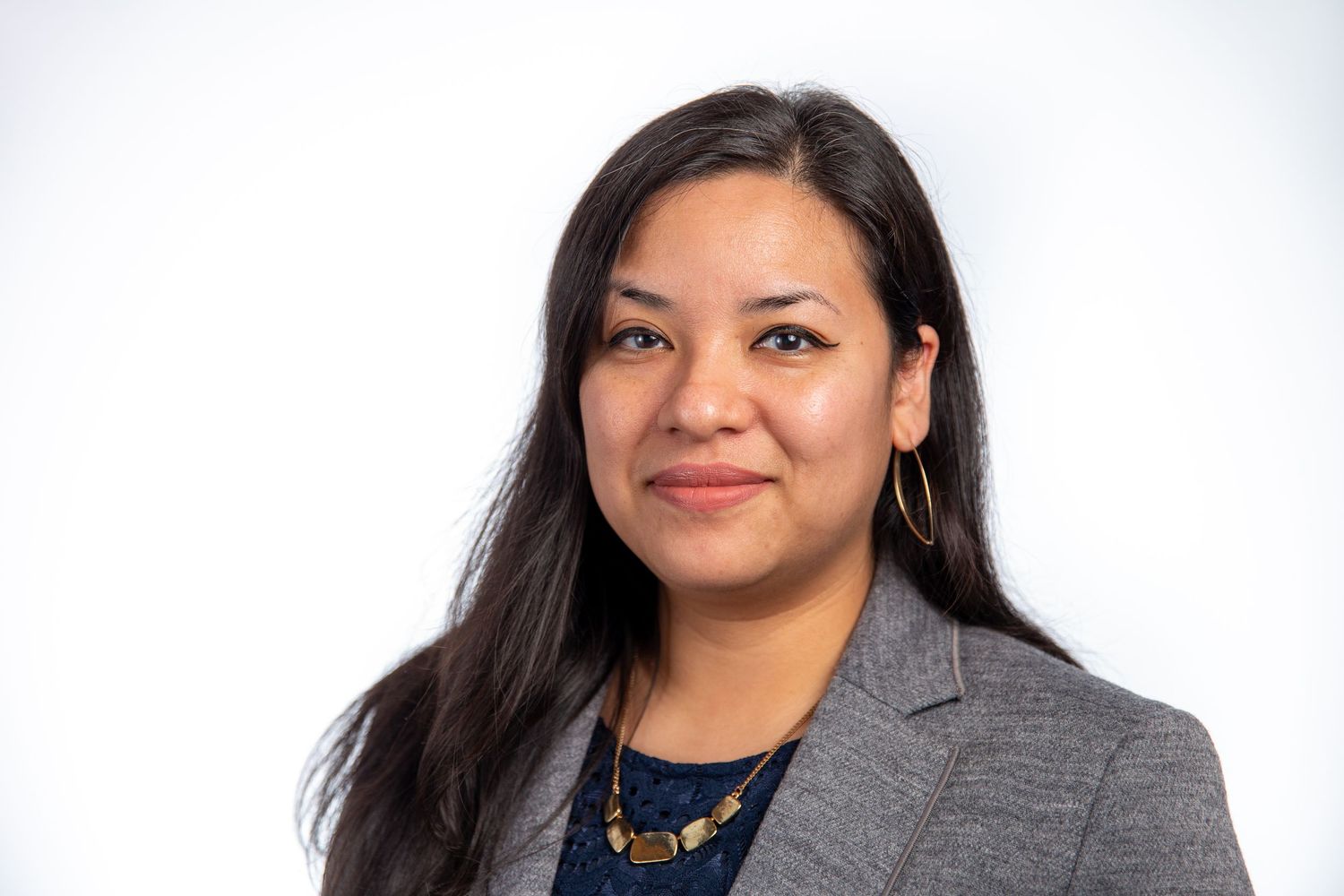 Professional woman with long dark hair wearing a gray blazer and gold necklace.
