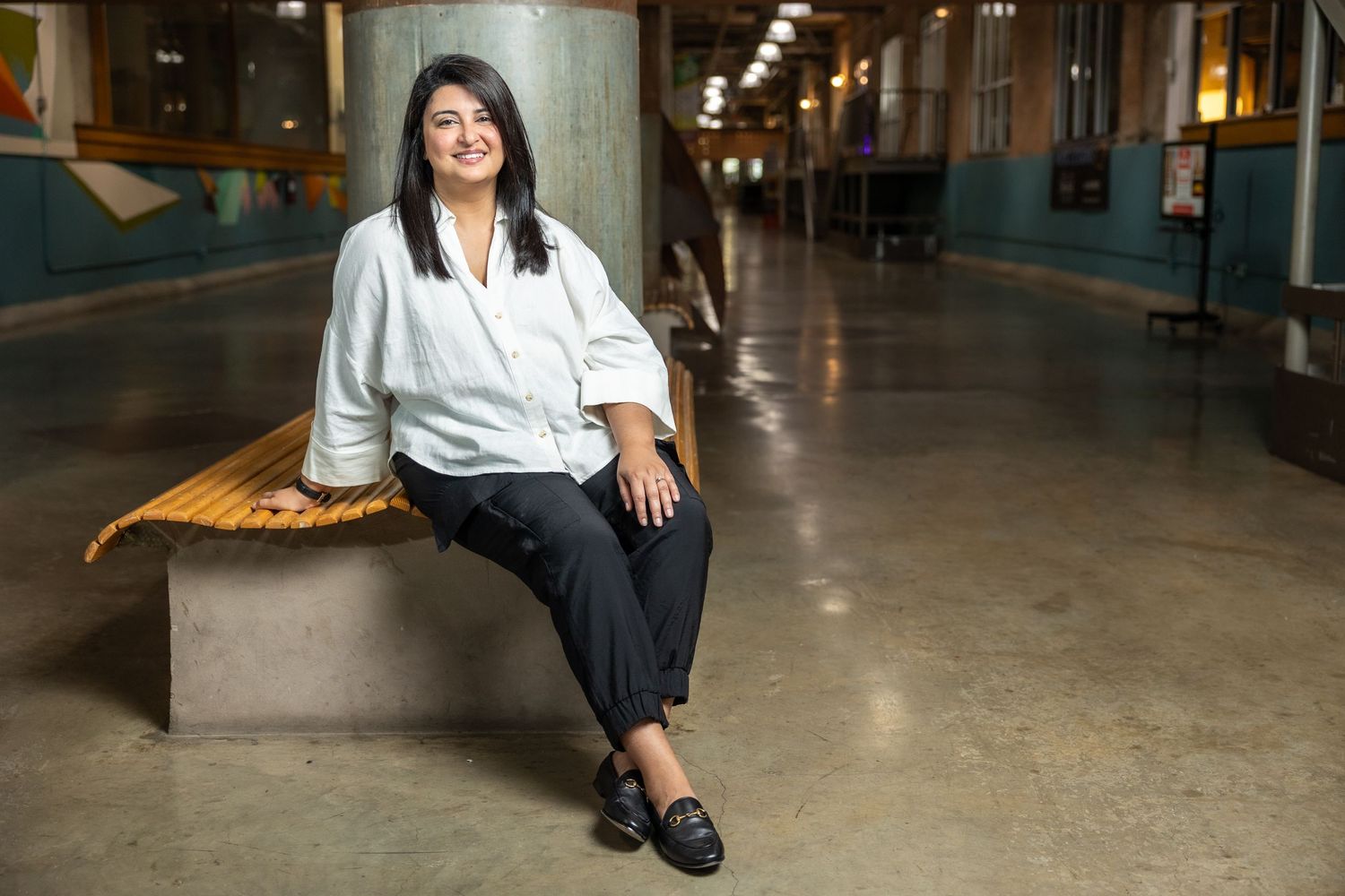 Smiling woman in white shirt and black pants sitting on bench in modern indoor hallway.