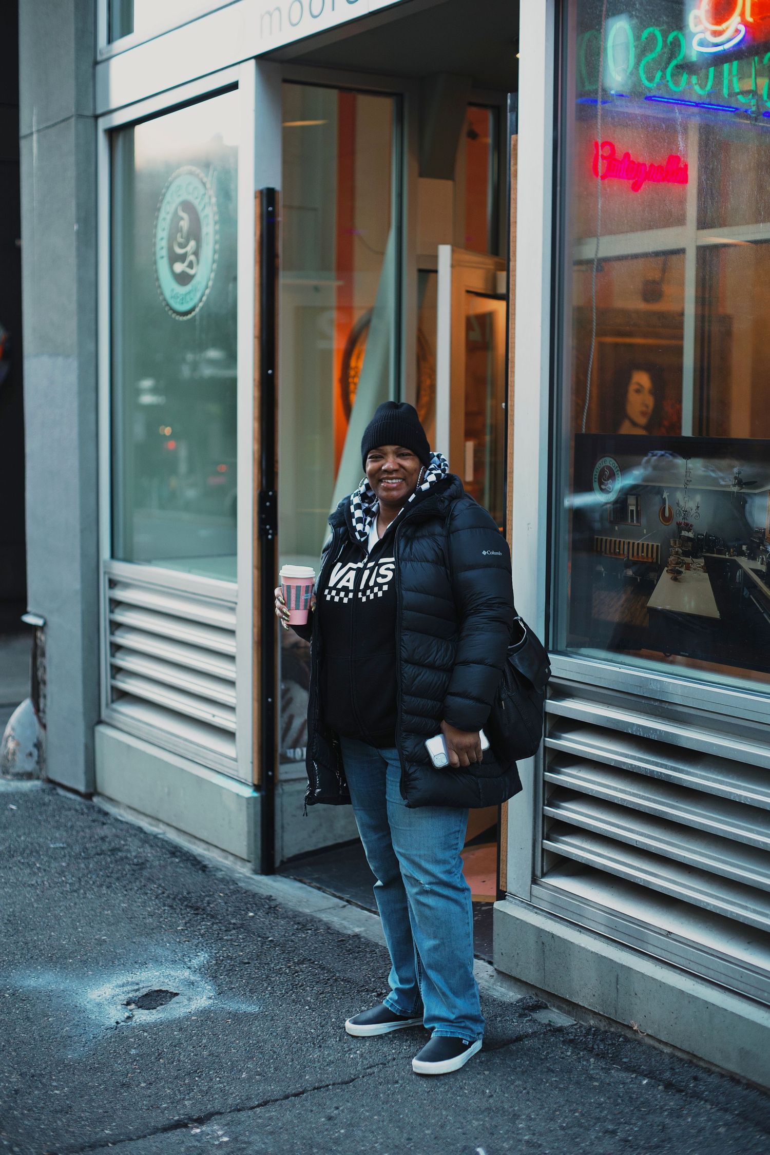 Person in black puffer jacket holding drink outside urban storefront.
