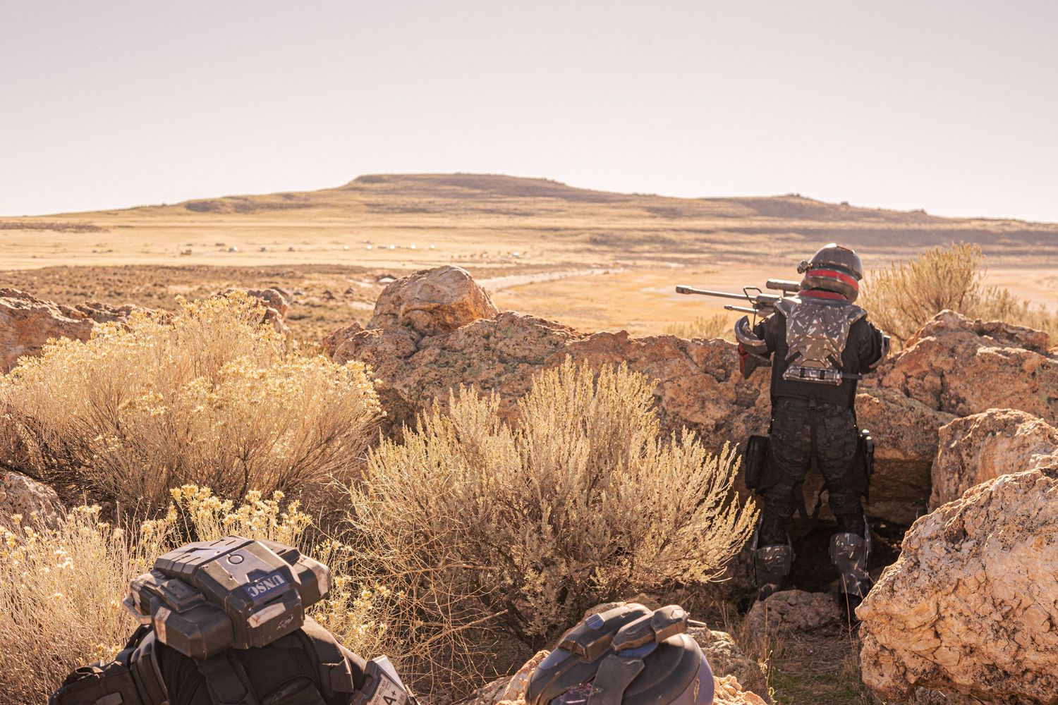Hunter in camouflage gear aiming rifle through desert sagebrush landscape with rocky terrain.