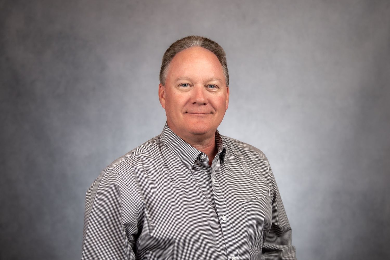 Middle-aged man with gray hair wearing a gray button-up shirt, professional headshot on gray background.