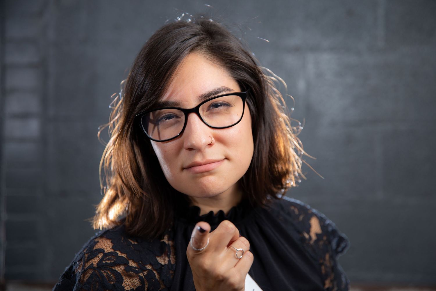 Young woman with dark hair and black glasses wearing a lace top, posing confidently against a gray wall.