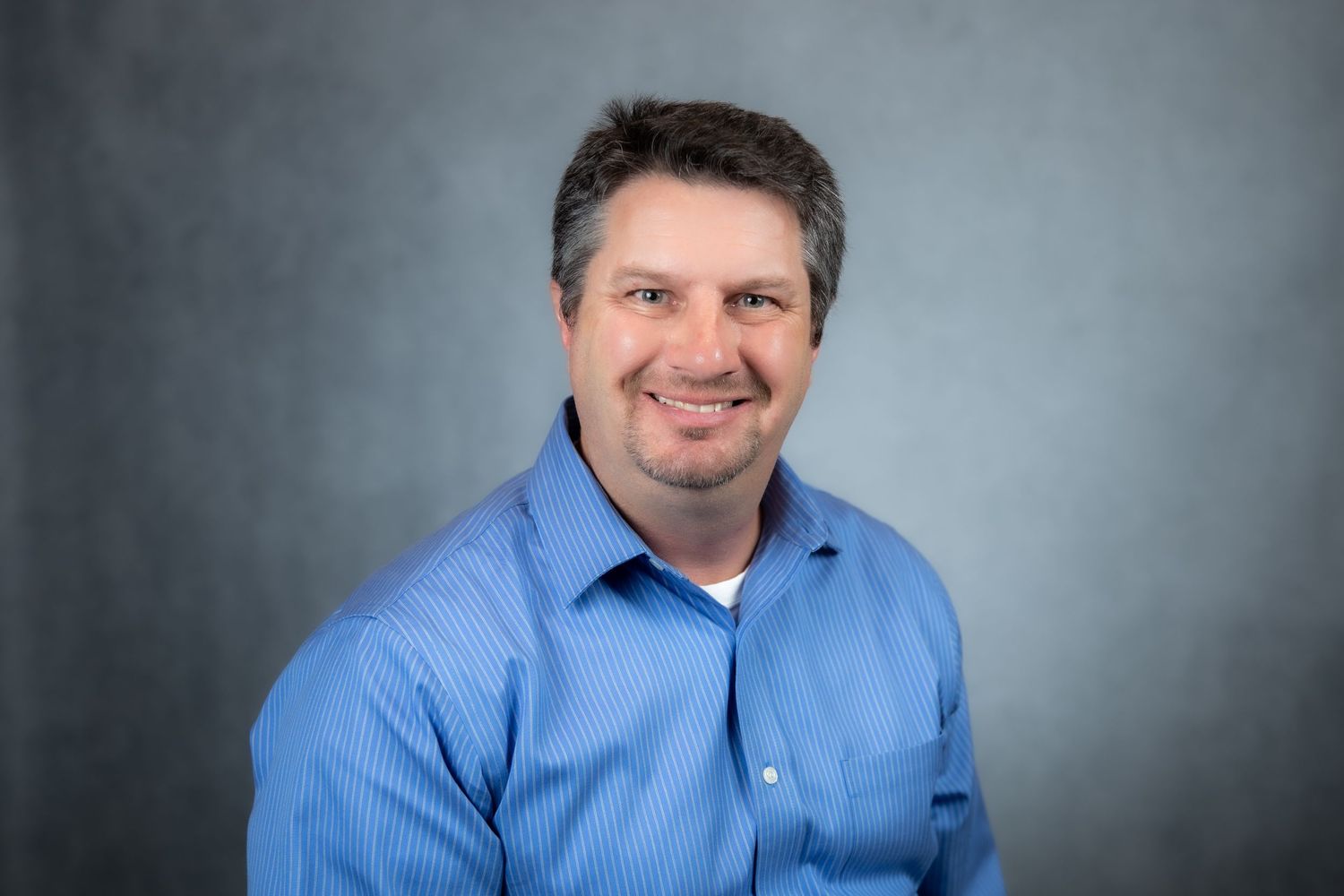 Smiling middle-aged man in a blue button-up shirt posing for a professional headshot on gray background.