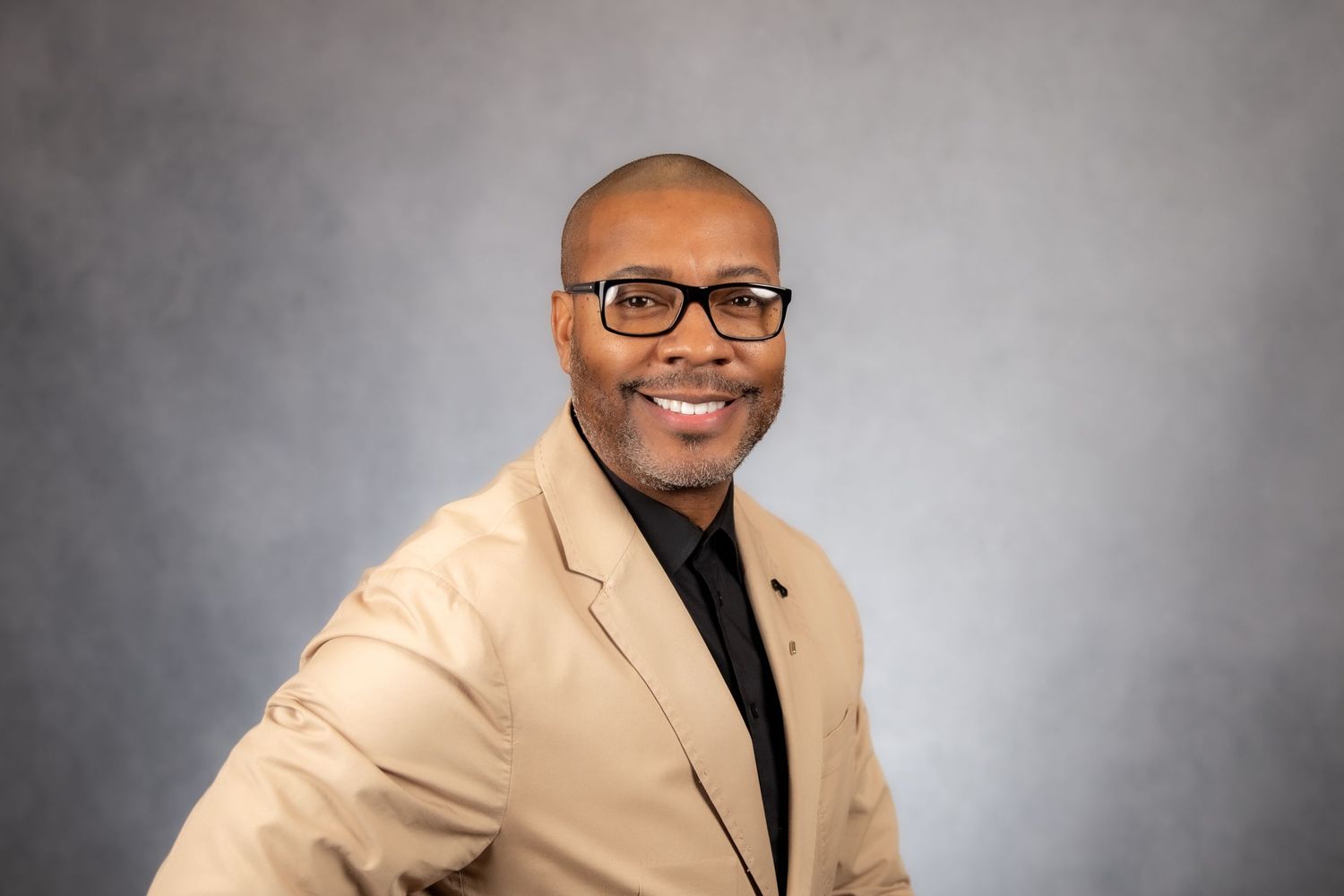 Smiling bald man wearing glasses and beige blazer with black shirt on gray background.